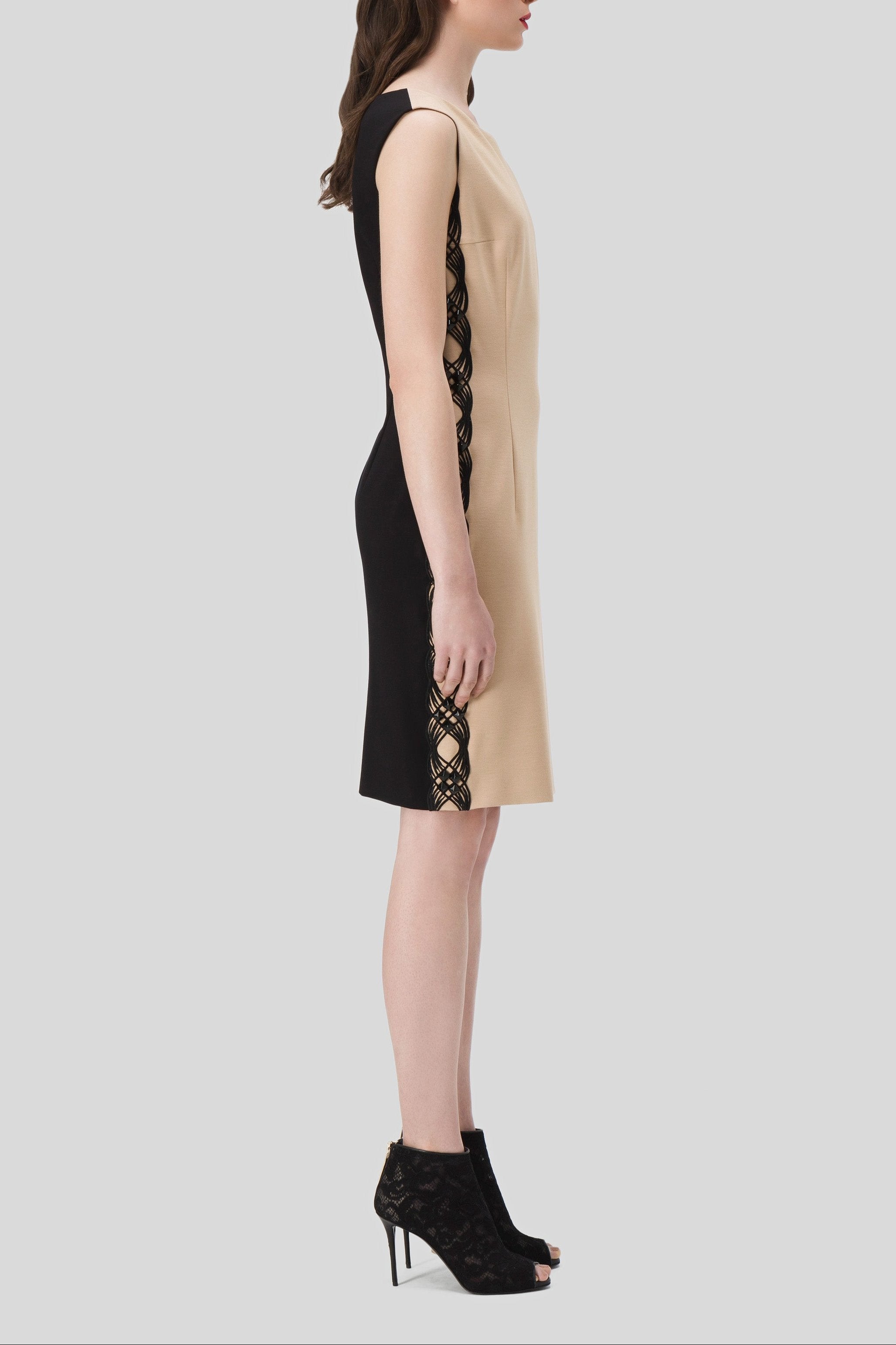 A woman in an Antonio Croce Adelin sleeveless dress featuring black and beige tones, with intricate side panel details, stands in profile view. She pairs the elegant dress with black high-heeled shoes against a plain gray background.