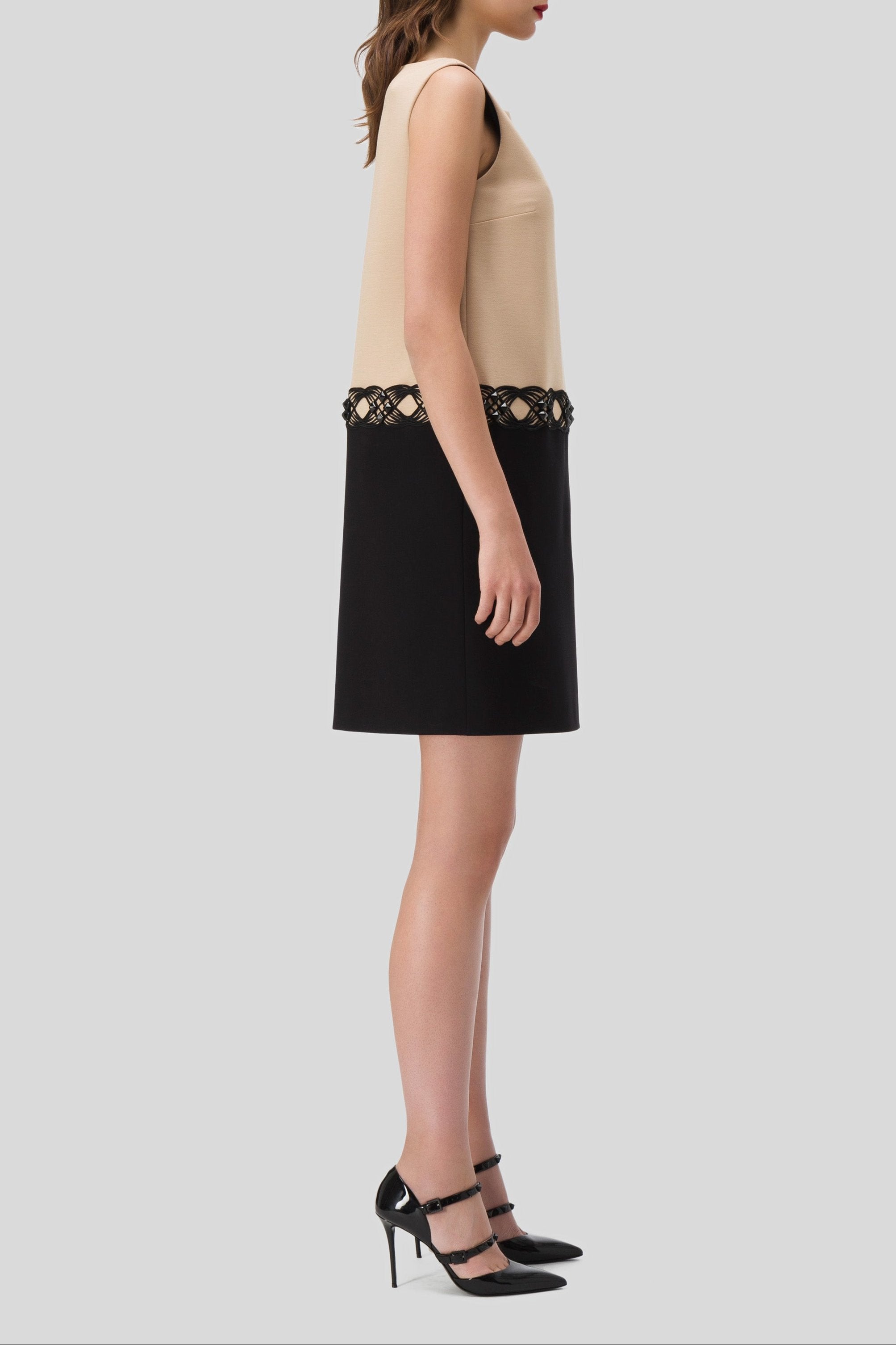 Side profile of a woman wearing the Antonio Croce Agave Two-Tone Contrast Dress, featuring a sleeveless beige design with decorative black lace trim, paired with a black skirt for a vintage-inspired look. She stands against a plain gray background while donning black high heels, her hair styled in soft waves.