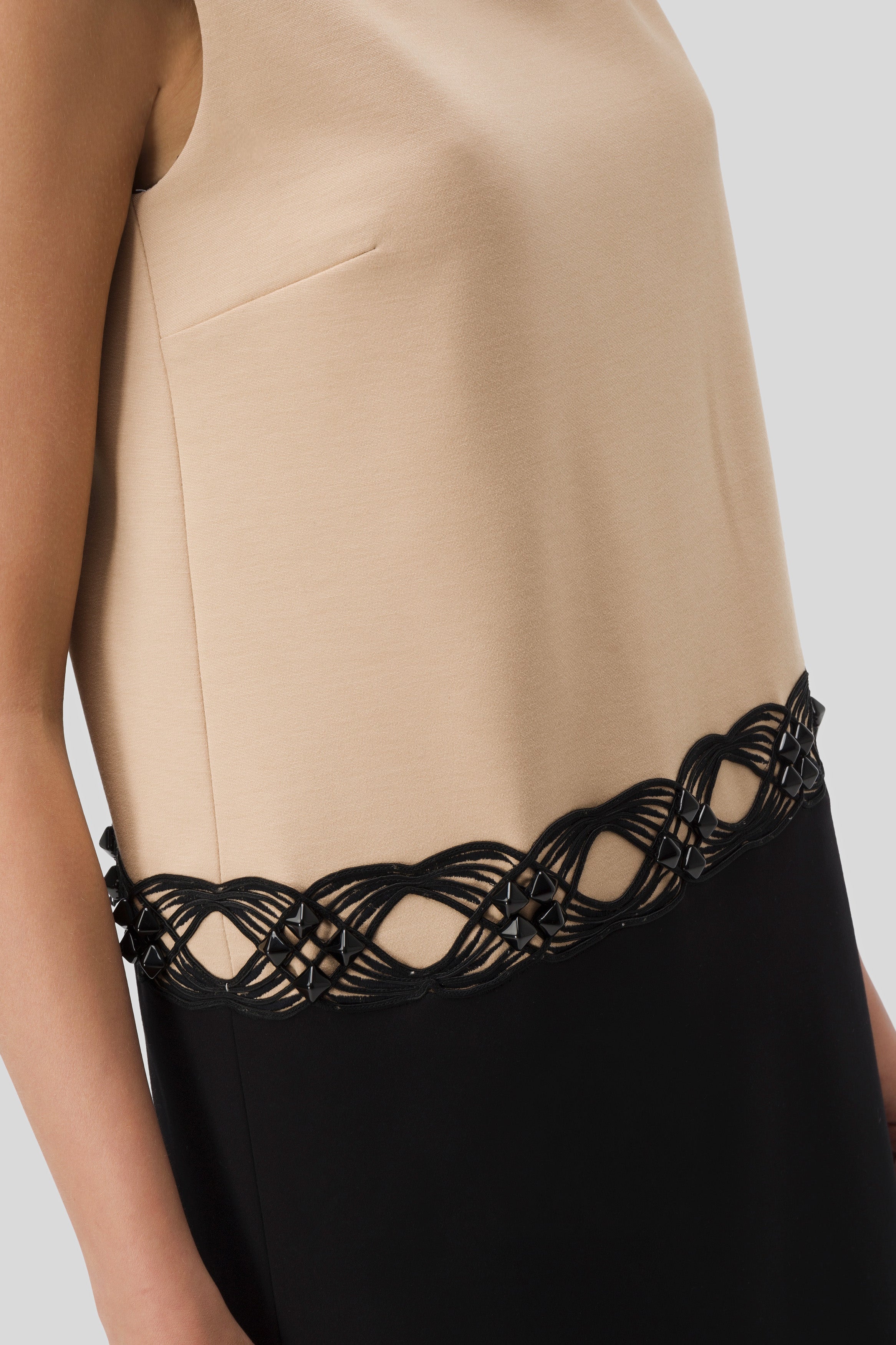 A close-up of the Antonio Croce Agave Two-Tone Contrast Dress showcases its sleeveless design with a beige top and black skirt. The elegant illusion neckline is beautifully complemented by a decorative black lattice trim featuring intricate, intertwined patterns, all set against a neutral background.