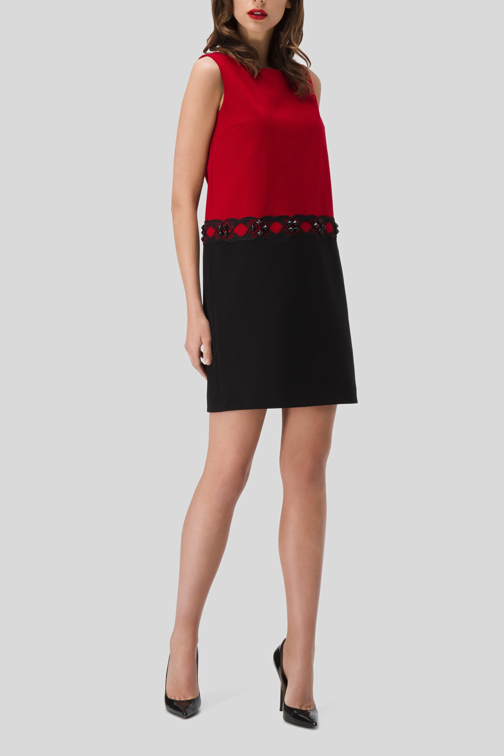 A woman wears the Antonio Croce Agave Two-Tone Contrast Dress, showcasing a sleeveless red top and black skirt with an illusion neckline and a decorative band at the waist. She stands against a plain gray background, exuding elegance in her black high heels.