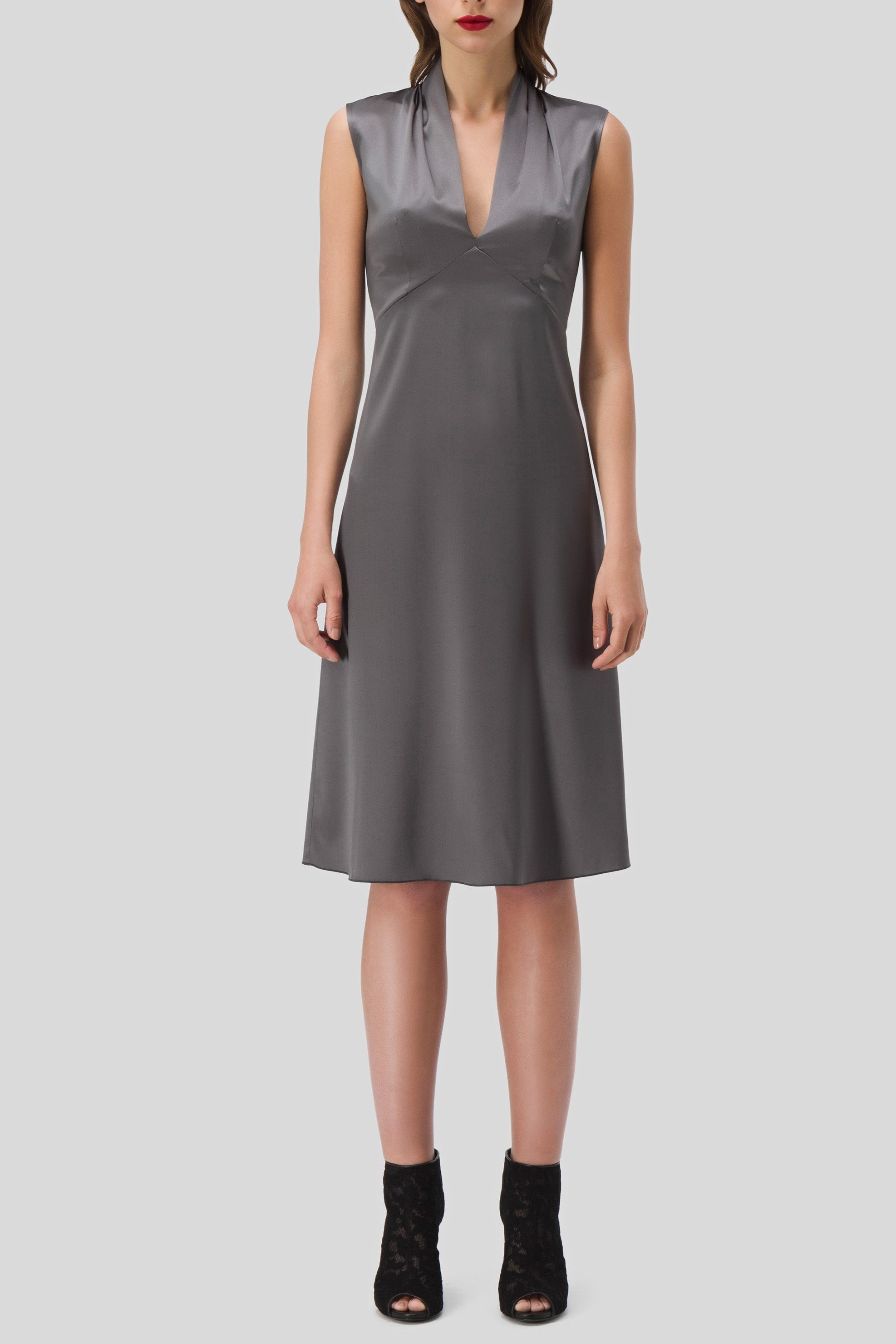 A person is wearing the Antonio Croce Akela Sleeveless Dress with Mesh Detail, featuring a knee-length design and deep V-neck in dark gray, standing against a plain backdrop. The minimalist style is complemented by black ankle boots, evoking the elegance of an Antonio Croce vintage piece.