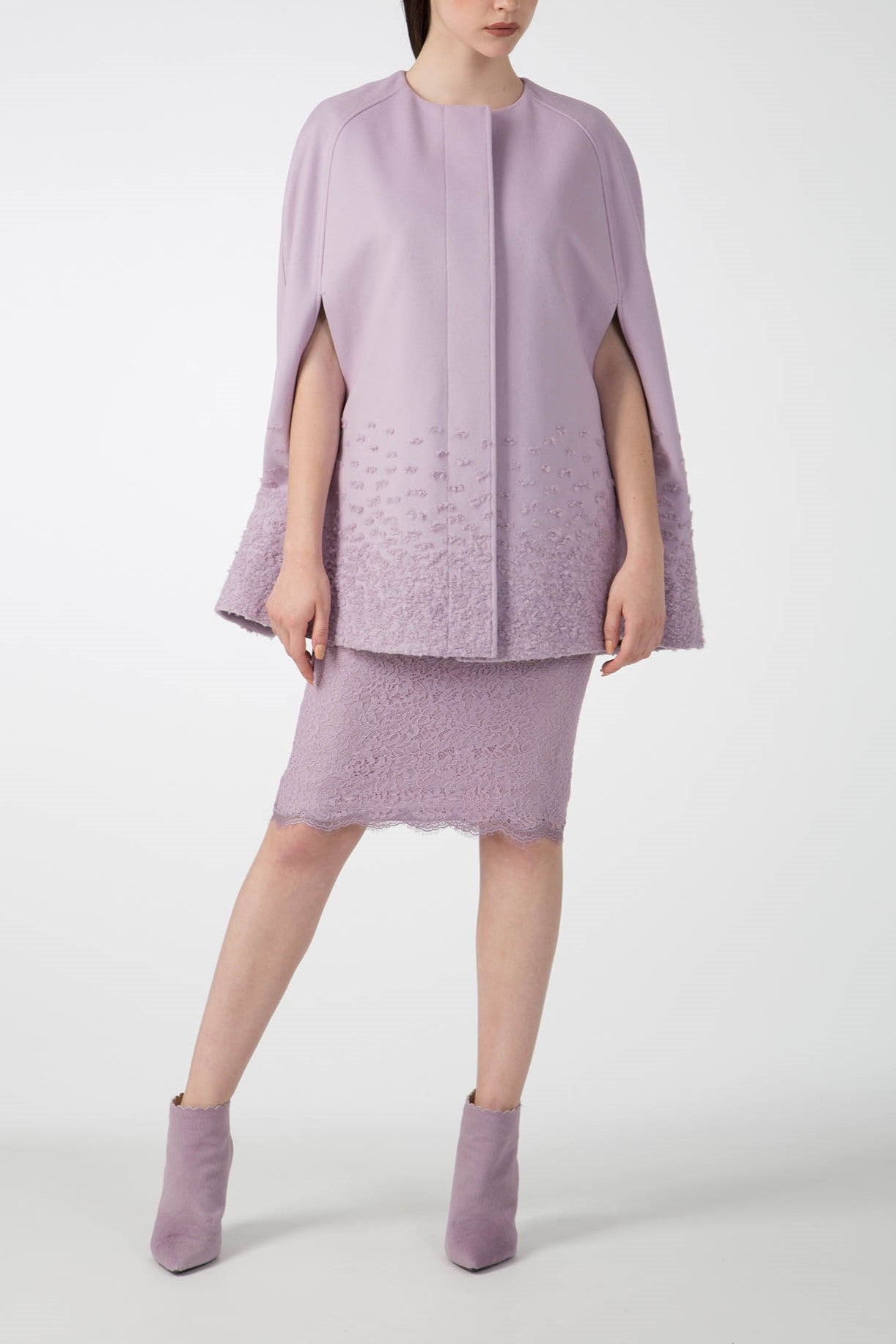 A model elegantly presents a vintage-inspired ensemble, featuring a lilac Antonio Croce Alex Cape Coat paired with a textured knee-length skirt made from luxurious woven fabric. The outfit is stylishly completed with lilac ankle boots against a plain white background.