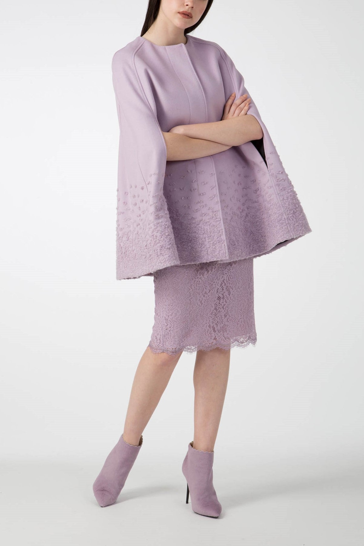 Standing confidently with arms crossed, a person is wrapped in the Antonio Croce Alex Cape Coat, made from luxurious light purple woven fabric. This stylish piece is elegantly paired with a matching lace skirt and completed with ankle-high purple boots, all set against a plain white backdrop.