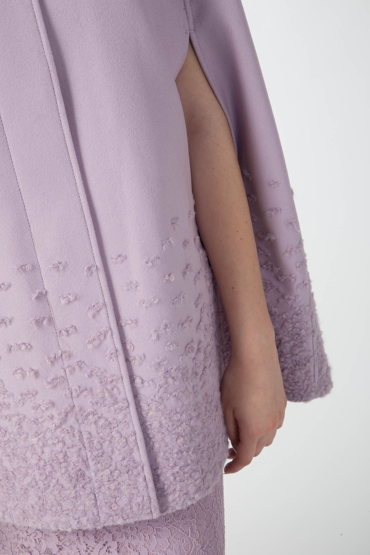 A close-up of the lavender Antonio Croce Alex Cape Coat highlights its luxurious woven fabric near the hem. The garment features a side slit that reveals part of the arm, offering a stylish contrast against its smooth upper section.