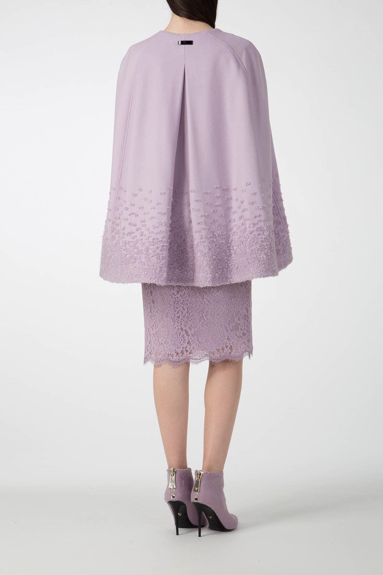 Wearing the luxurious Antonio Croce Alex Cape Coat, a woman in a lavender dress with lace details stands facing away. She complements her vintage-inspired ensemble with ankle-high, lavender high-heeled boots featuring zippers. The plain white background emphasizes the elegance of the woven fabric by Antonio Croce.