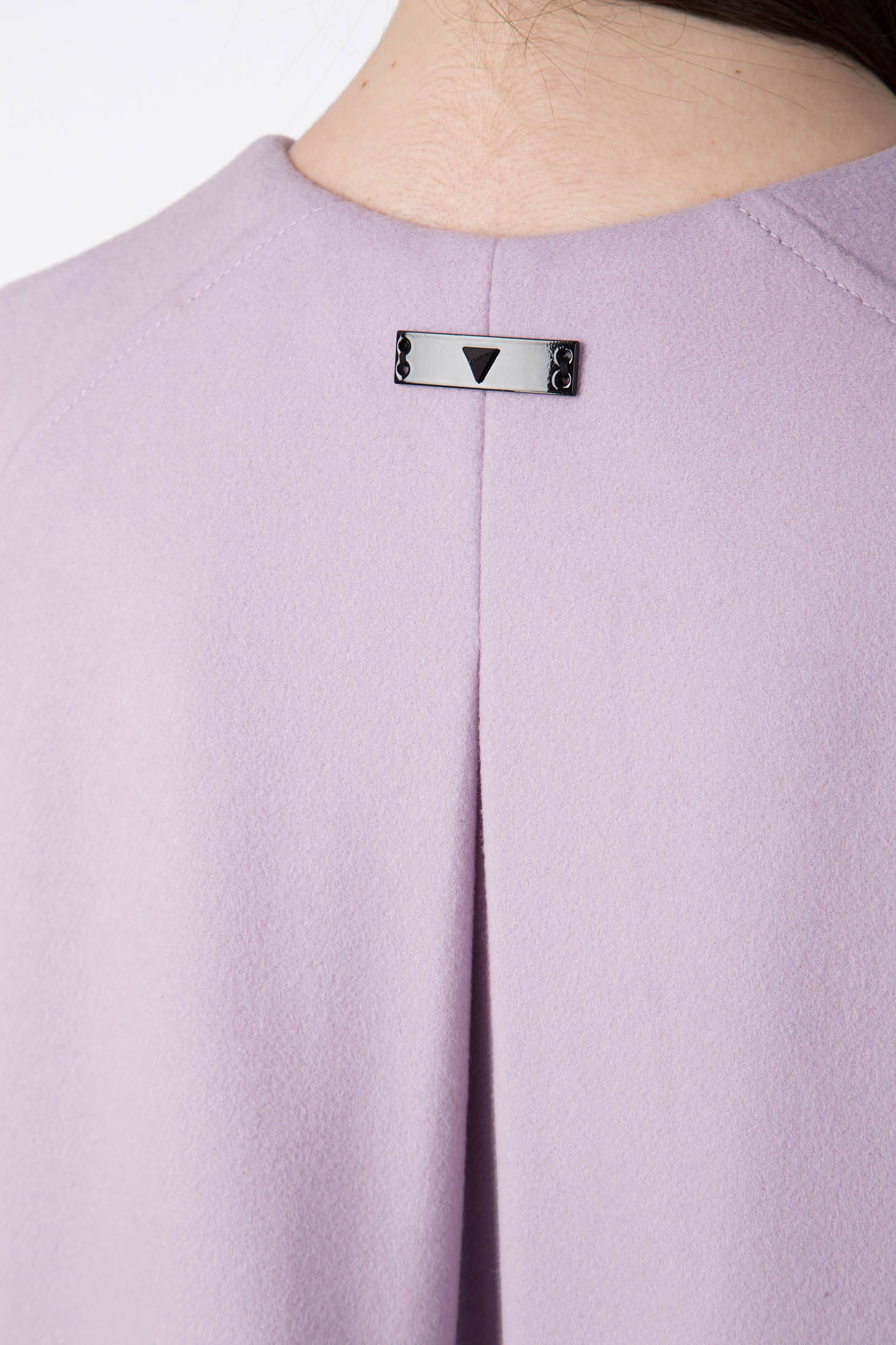 Close-up of a person wearing the Antonio Croce Alex Cape Coat in lavender. The back features a small, rectangular metal plate with a black triangle detail, adding a vintage touch. The luxurious woven fabric includes a central pleat beneath the tag.