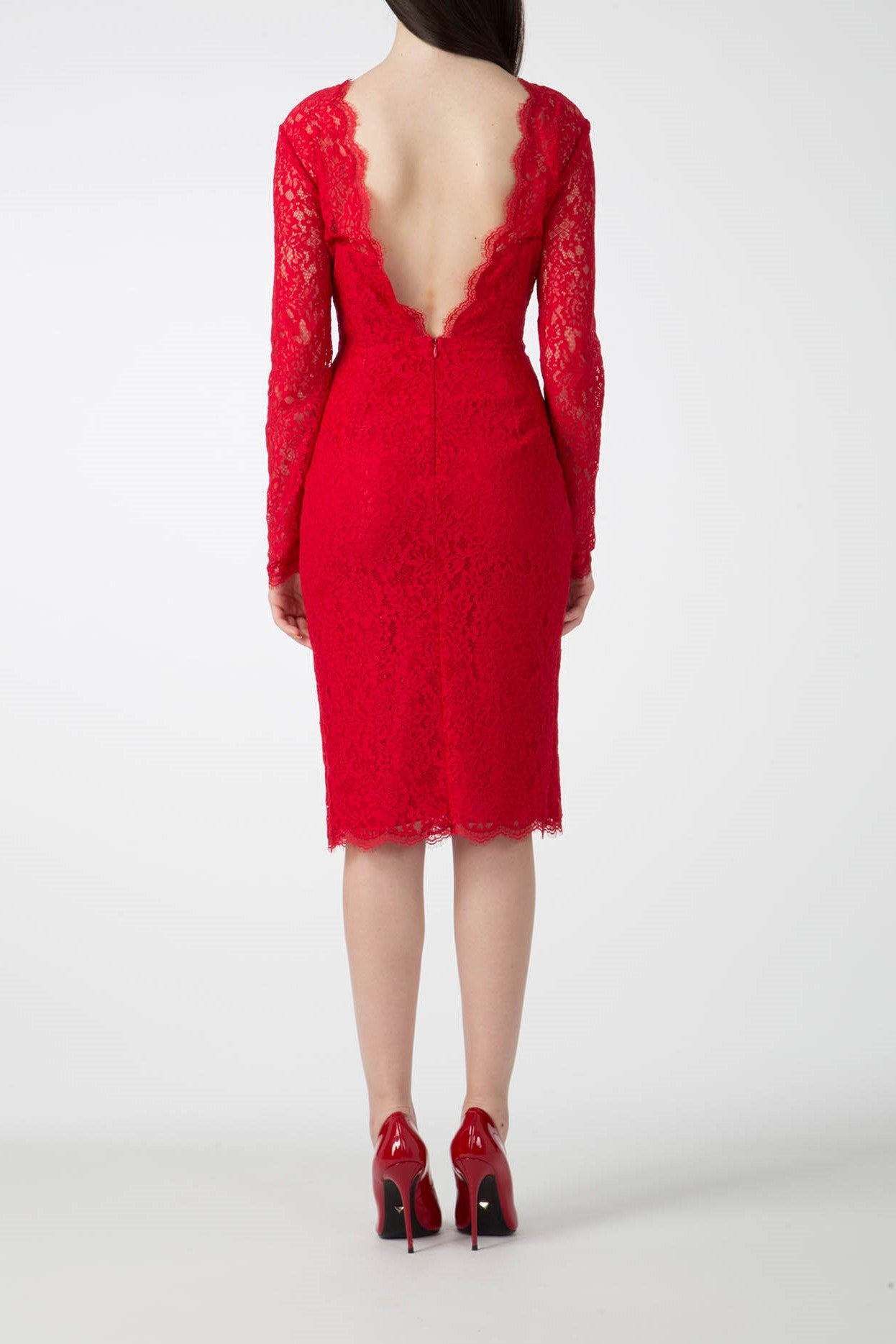 A woman in an Antonio Croce Ambeta Long-Sleeve Lace Dress, featuring a vintage design and deep V-back, paired with red high heels, stands facing away. Her long, dark hair cascades straight down her back. The elegant Antonio Croce dress boasts long sleeves and falls just below the knee, enhancing her sophisticated look.