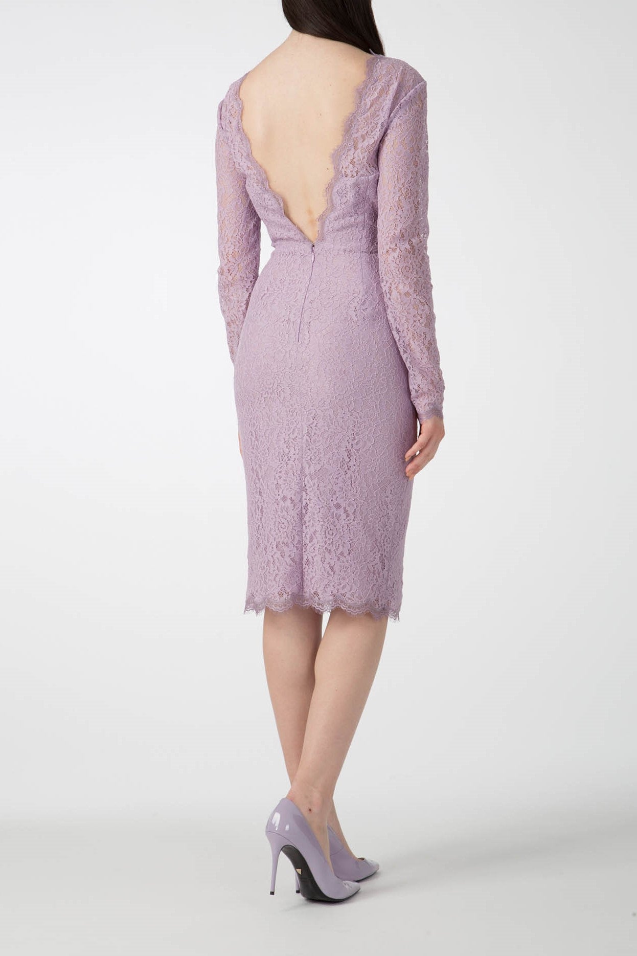 The woman exudes elegance in the Antonio Croce Ambeta Long-Sleeve Lace Dress, showcasing its deep V-back and vintage charm as she stands facing away. The lilac dress, with its intricate lace design, features long sleeves and a knee-length hem. She complements her look with matching lilac high-heeled shoes against a plain white background.