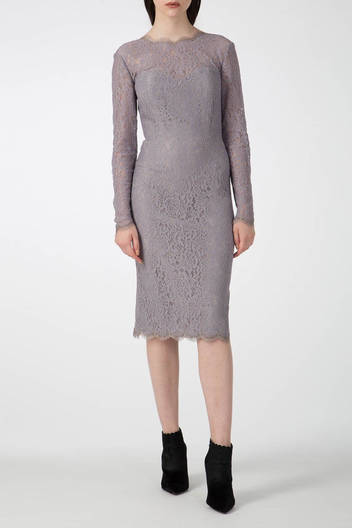 A woman wearing the Antonio Croce Ambeta Long-Sleeve Lace Dress, which features a form-fitting design and light gray lace fabric with a knee-length hem and scalloped edges, stands against a plain background. This vintage piece exudes an elegant look, perfectly complemented by her black ankle boots.