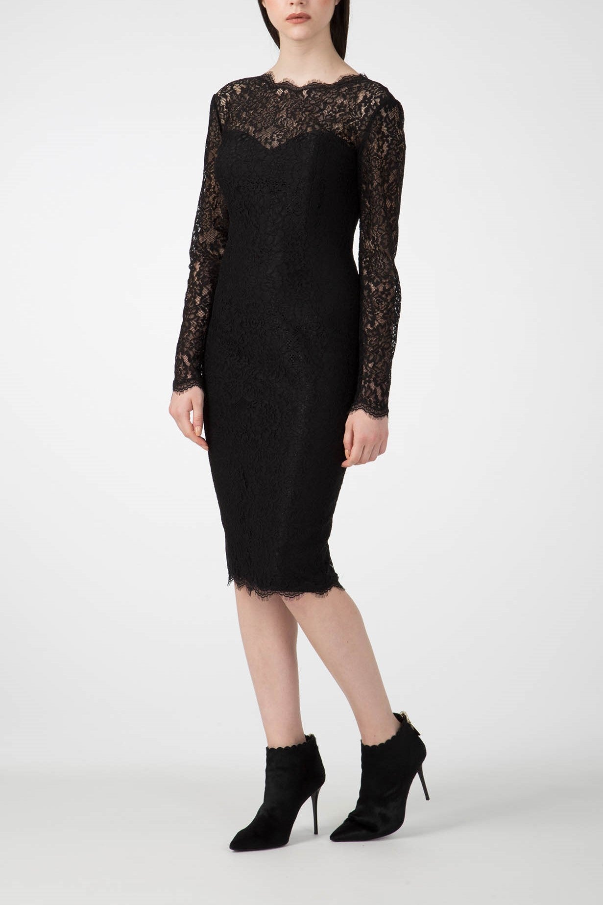 Wearing the Antonio Croce Ambeta Long-Sleeve Lace Dress, a person exudes elegance as they stand against a plain background. The dress, known for its fitted silhouette and sheer lace detail at the top, features long sleeves and a knee-length hem that enhance its vintage allure, perfectly complemented by black high-heeled ankle boots.