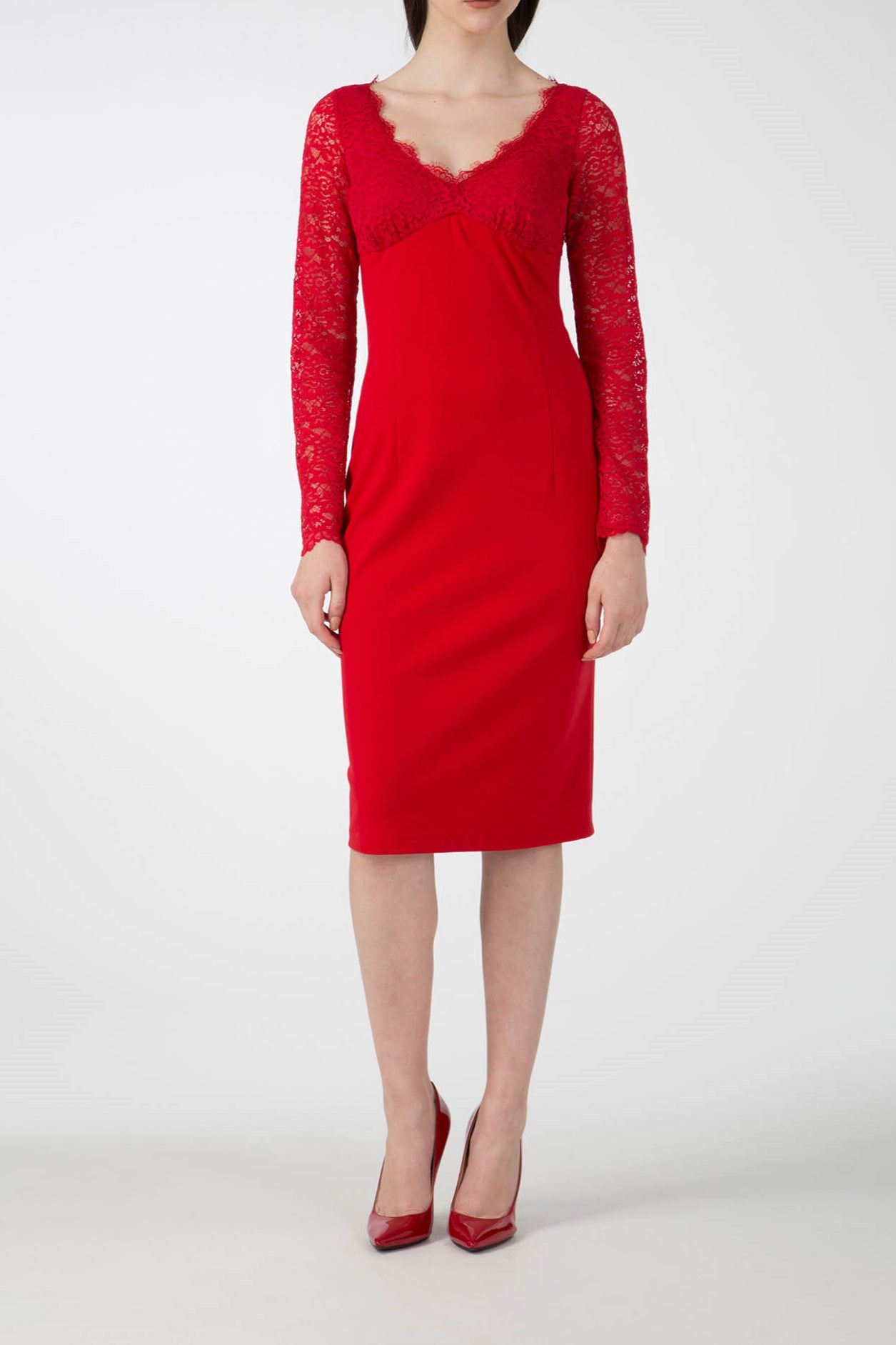 A woman wearing the Antonio Croce Ampara Embroidered Lace Dress, featuring an intricate design and delicate lace sleeves, stands against a plain white background. She complements the knee-length, fitted silhouette with matching red high-heeled shoes. Her hands rest elegantly at her sides.