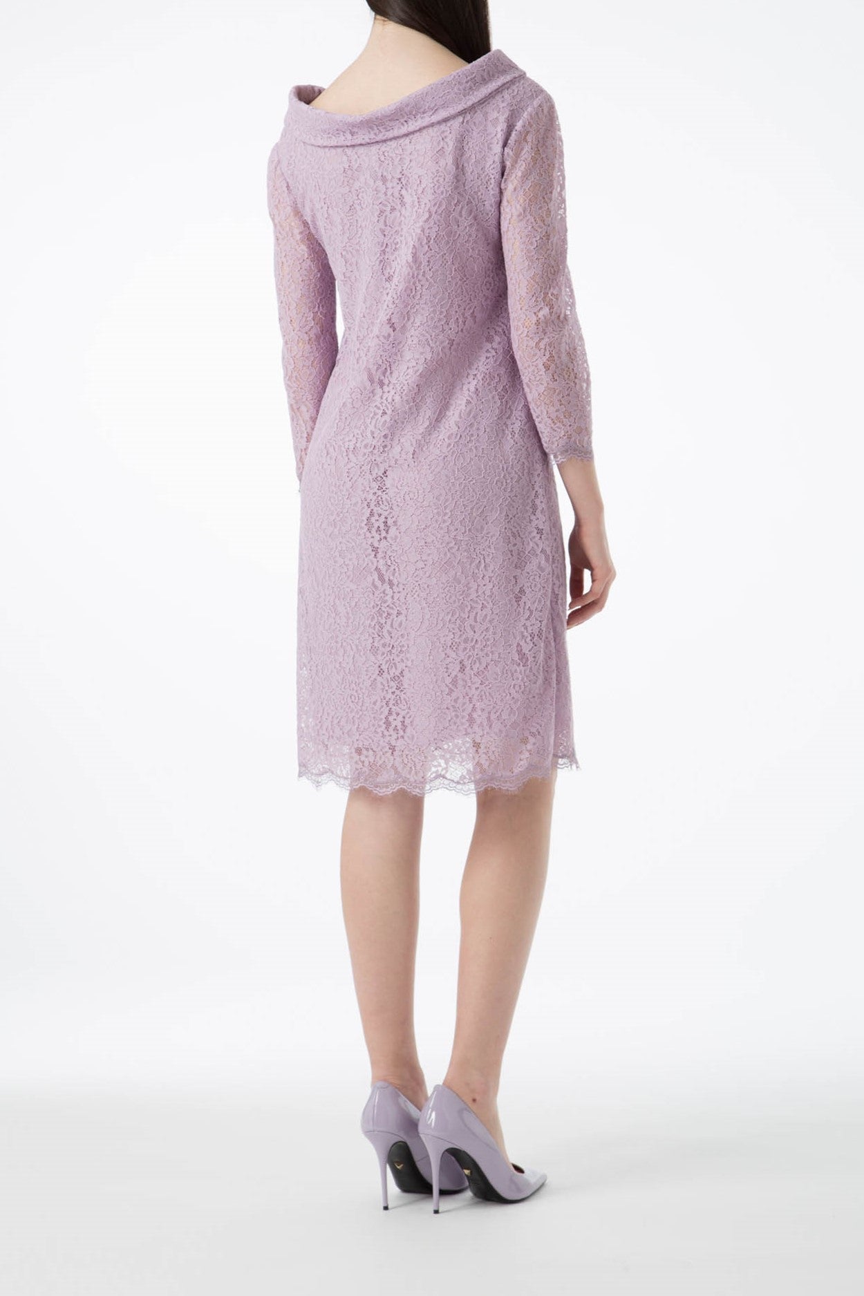 A woman wearing the Antonio Croce Ana Romantic Lace Dress, a vintage knee-length piece in lavender lace, stands with her back to the camera. This elegant design showcases three-quarter sleeves and a draped neckline, paired perfectly with matching lavender high heels against a plain white backdrop.