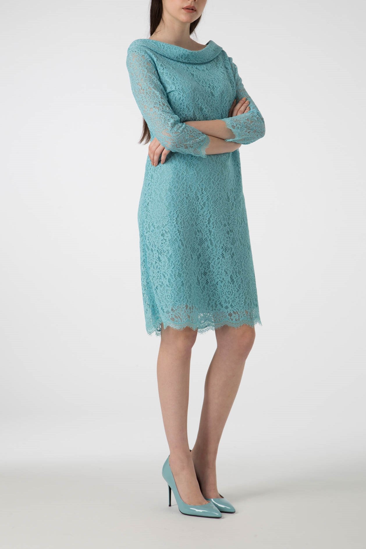 A person stands with arms crossed wearing the Antonio Croce Ana Romantic Lace Dress, a vintage, knee-length light blue lace dress paired with matching high heels. This elegant outfit features three-quarter sleeves and a scalloped hem, perfectly capturing timeless style. The background is plain white.