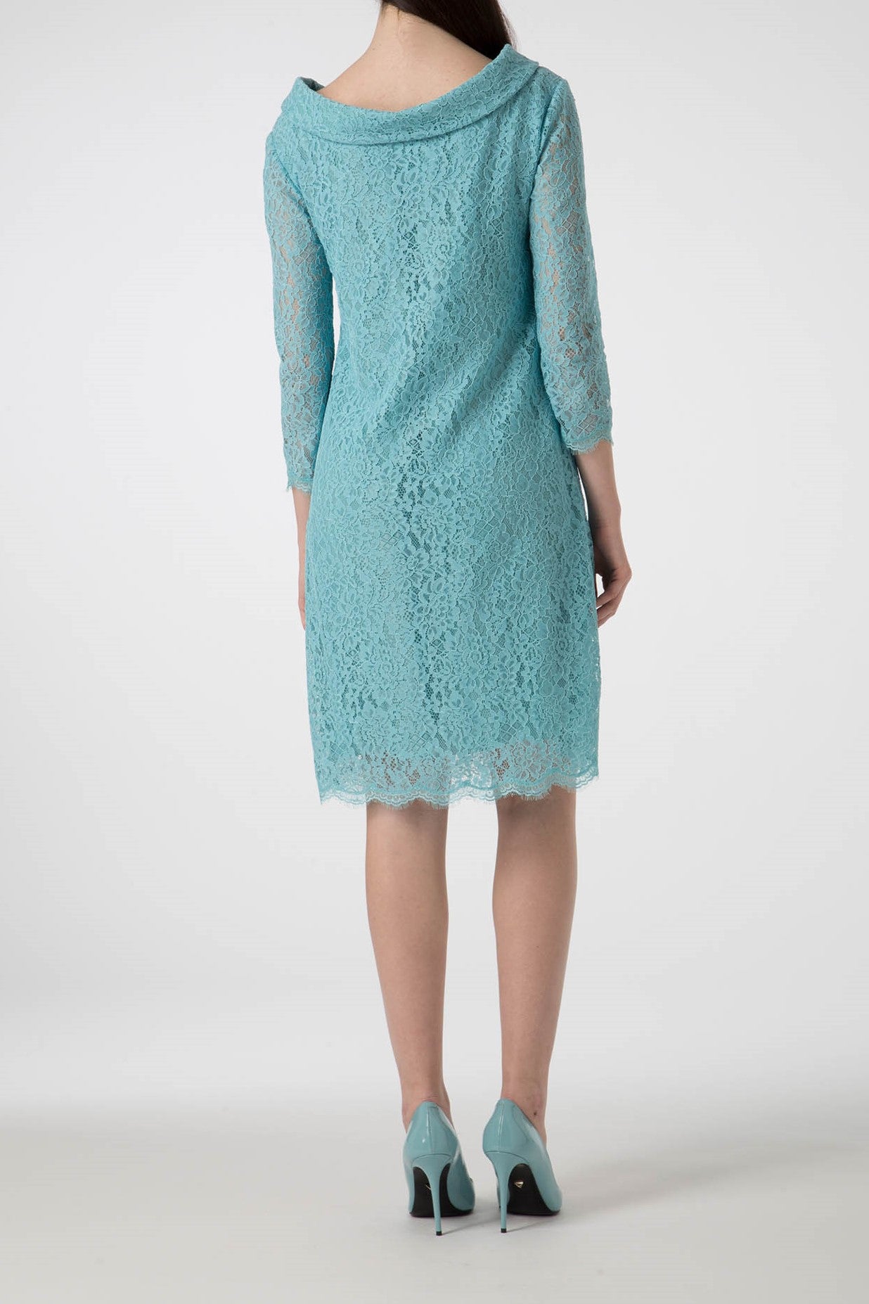 A person is wearing the Antonio Croce Ana Romantic Lace Dress, a vintage-style, knee-length dress with long sleeves in teal lace. They are also wearing matching teal high heels. The background is plain and light-colored as they stand with their back to the camera.