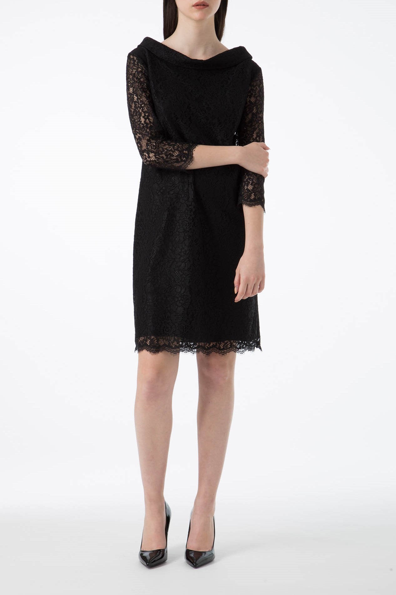 A woman wearing an Antonio Croce Ana Romantic Lace Dress, a knee-length vintage black lace dress with three-quarter sleeves, stands against a white background. She is also wearing black pointed-toe heels. Her arms are crossed, and her head is slightly turned to one side.