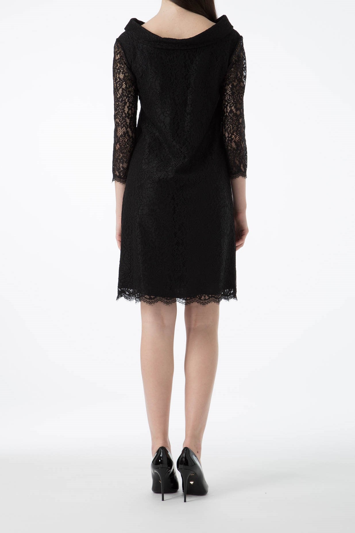 A woman in an Antonio Croce Ana Romantic Lace Dress, characterized by its vintage black lace and three-quarter sleeves, stands facing away from the camera. The elegant dress falls just above the knees and is elegantly complemented by black high heels against a plain white background.