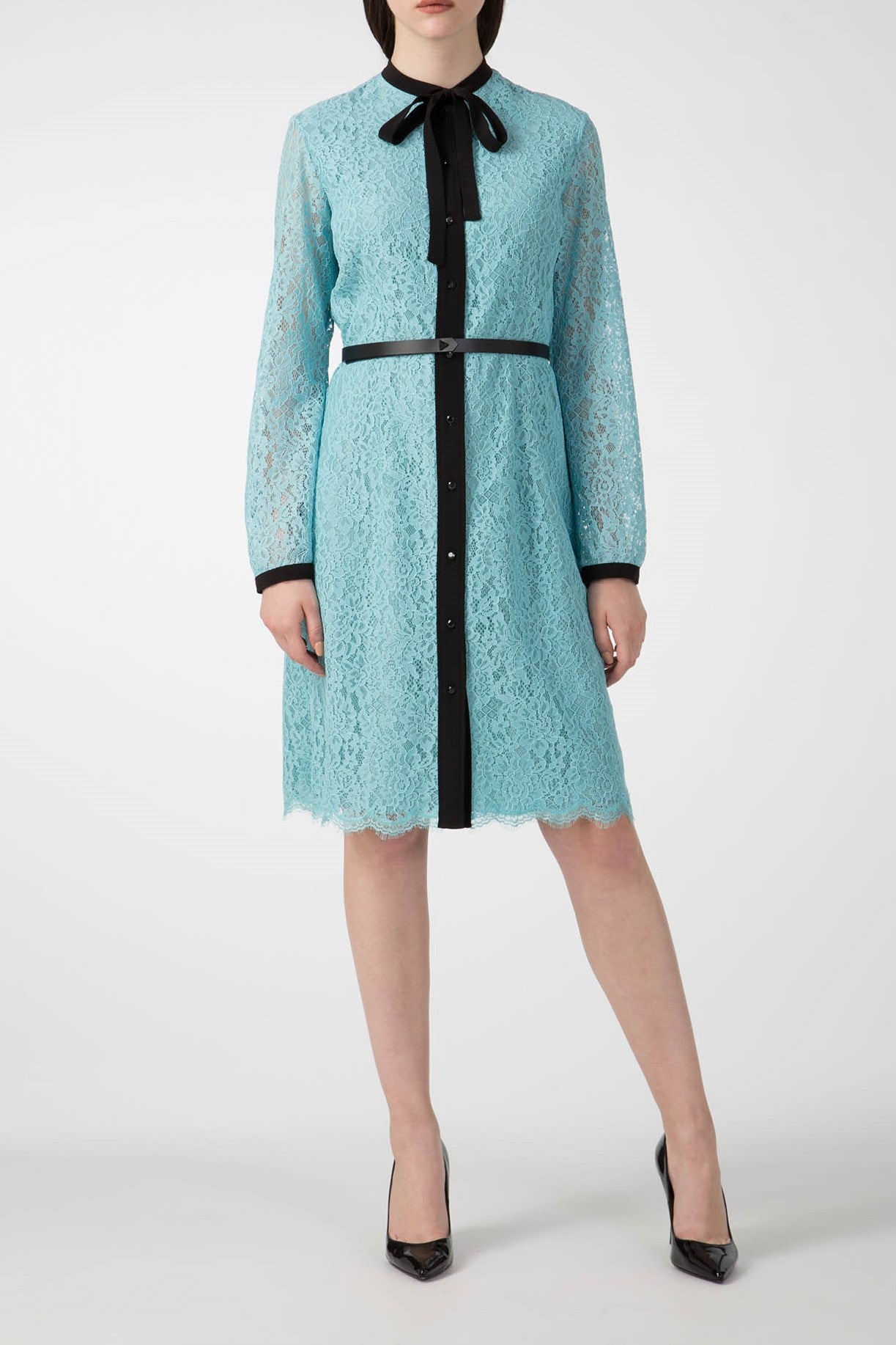 A person dressed in an Antonio Croce Anis Lace Shirt Dress with Bow, showcasing its unique sky blue lace design, long sleeves, and shirt collar adorned with a black bow. The dress features a distinctive black vertical panel with buttons, complemented by a matching black belt, and is paired with vintage items such as classic black heels.