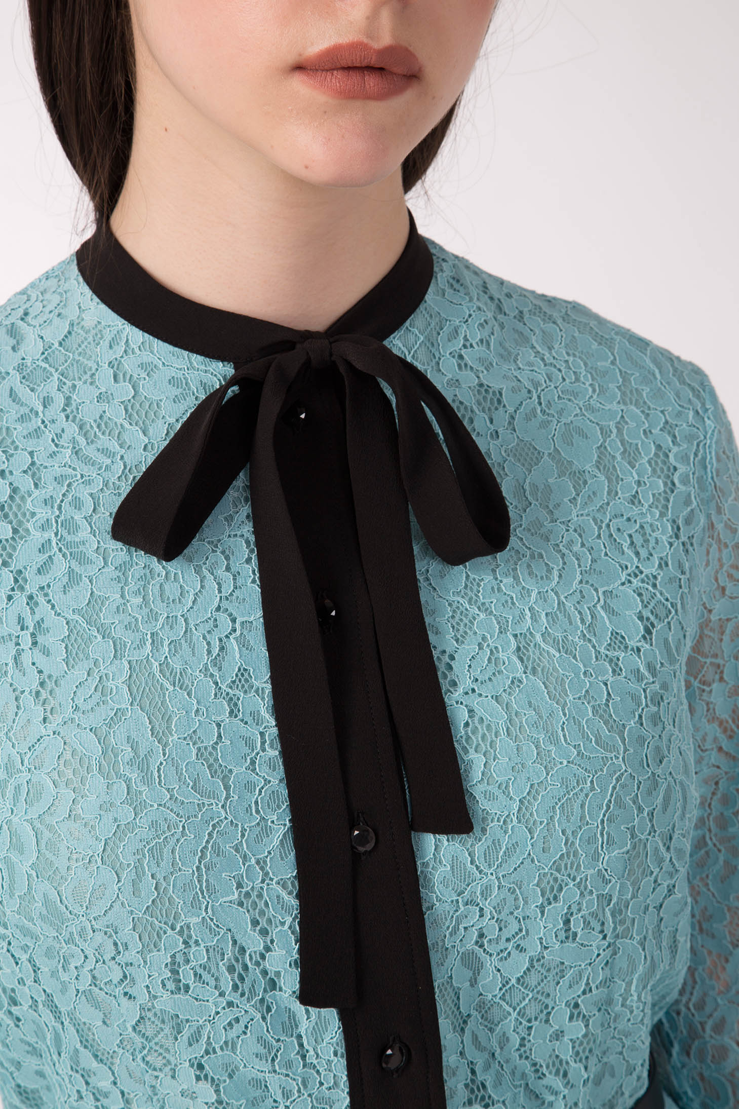 A person is seen wearing the Antonio Croce Anis Lace Shirt Dress with Bow, which is a vintage light blue lace piece featuring a black bow tie at the collar and black buttons down the front. The photo captures the lower half of their face and part of their upper body against a plain background.