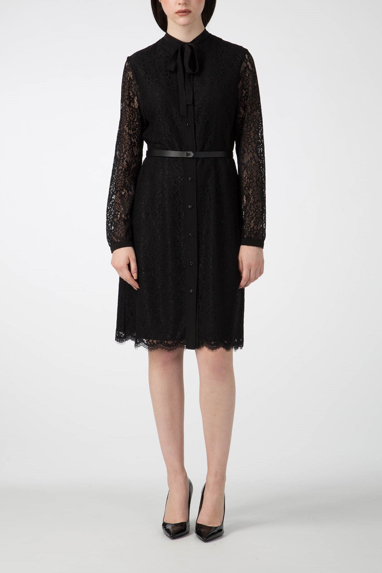 A person is wearing the Antonio Croce Anis Lace Shirt Dress with Bow, a vintage-inspired piece featuring long sleeves and a shirt collar adorned with a bow tie. The knee-length dress, designed by Antonio Croce, is buttoned down the front and features a thin black belt at the waist. They pair it perfectly with black high-heeled shoes against a plain white background.