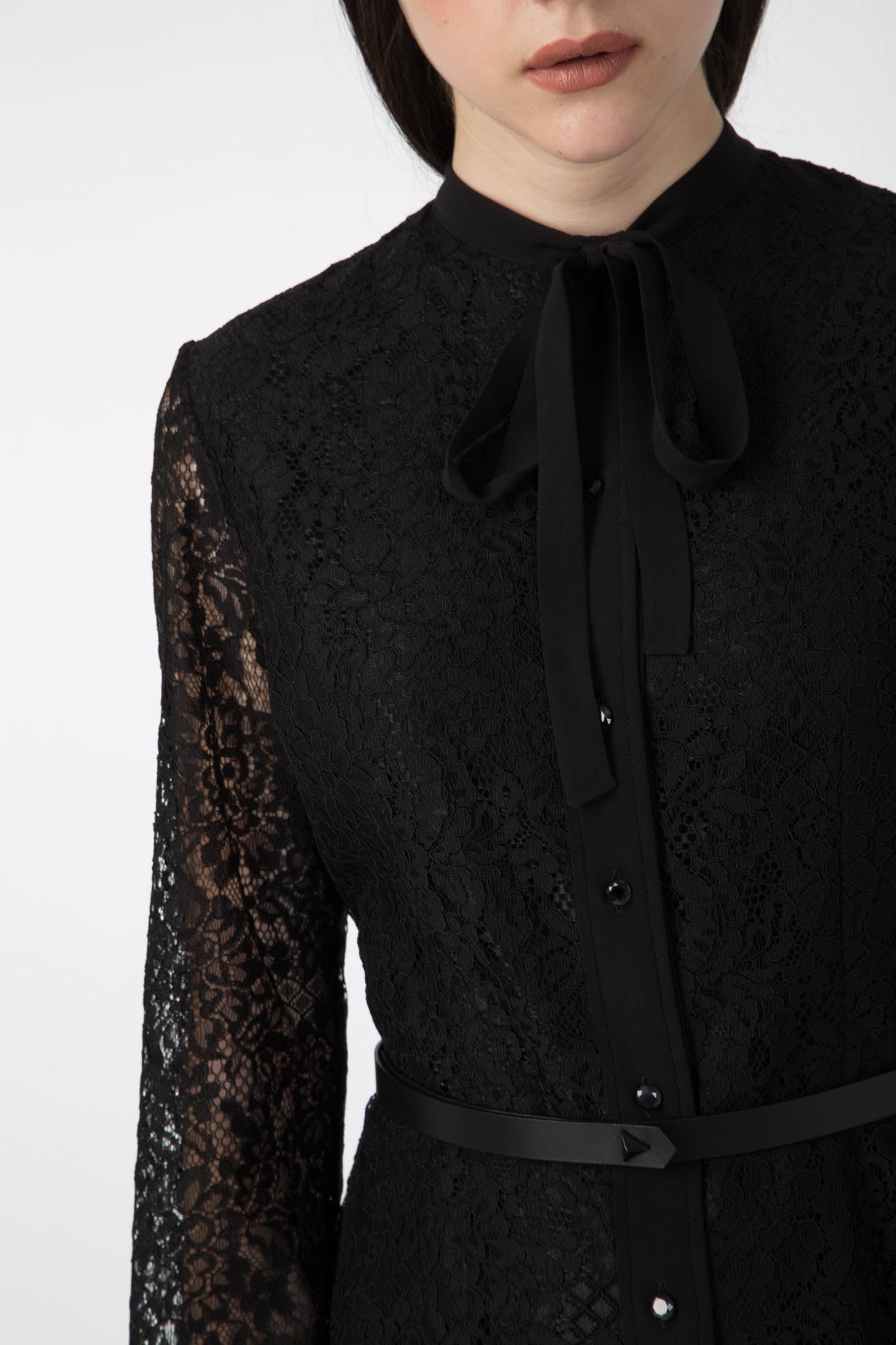 A woman stands gracefully in the Antonio Croce Anis Lace Shirt Dress, a vintage piece featuring a charming bow at the collar. The elegant black dress by Antonio Croce showcases long sleeves and delicate button details down the front, set against a plain white background.