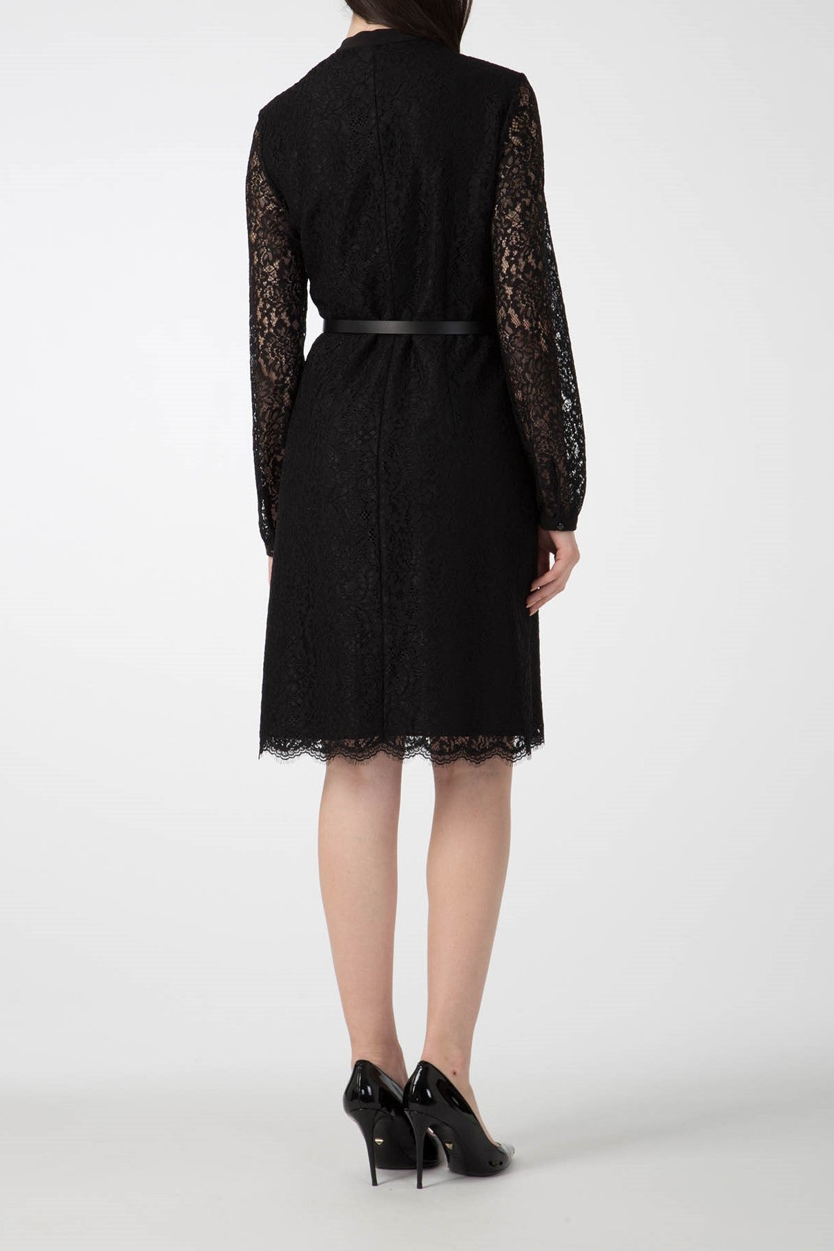 Wearing the Antonio Croce Anis Lace Shirt Dress with Bow, a person presents their back to the camera, showcasing its vintage lace design, long sleeves, and sleek black belt. The knee-length hem pairs perfectly with their black patent leather high heels. This ensemble effortlessly exudes a delicate blend of timeless elegance against a plain white background.