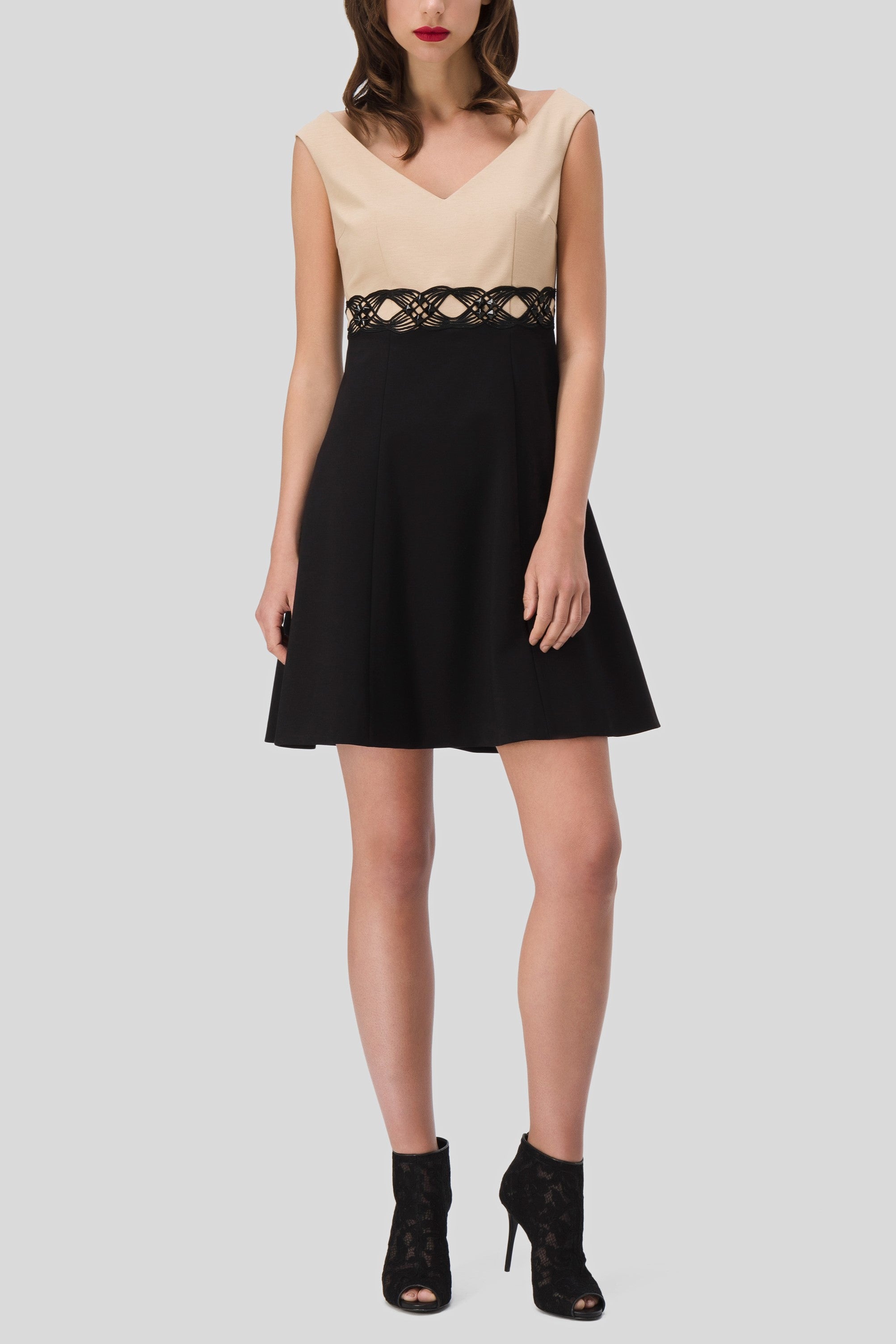 A woman in a sleeveless Antonio Croce Annely Dress with Layered Design, which is a timeless piece featuring a beige top, black skirt, and lace detail at the waist. She pairs it with black lace ankle boots against a plain light gray background.