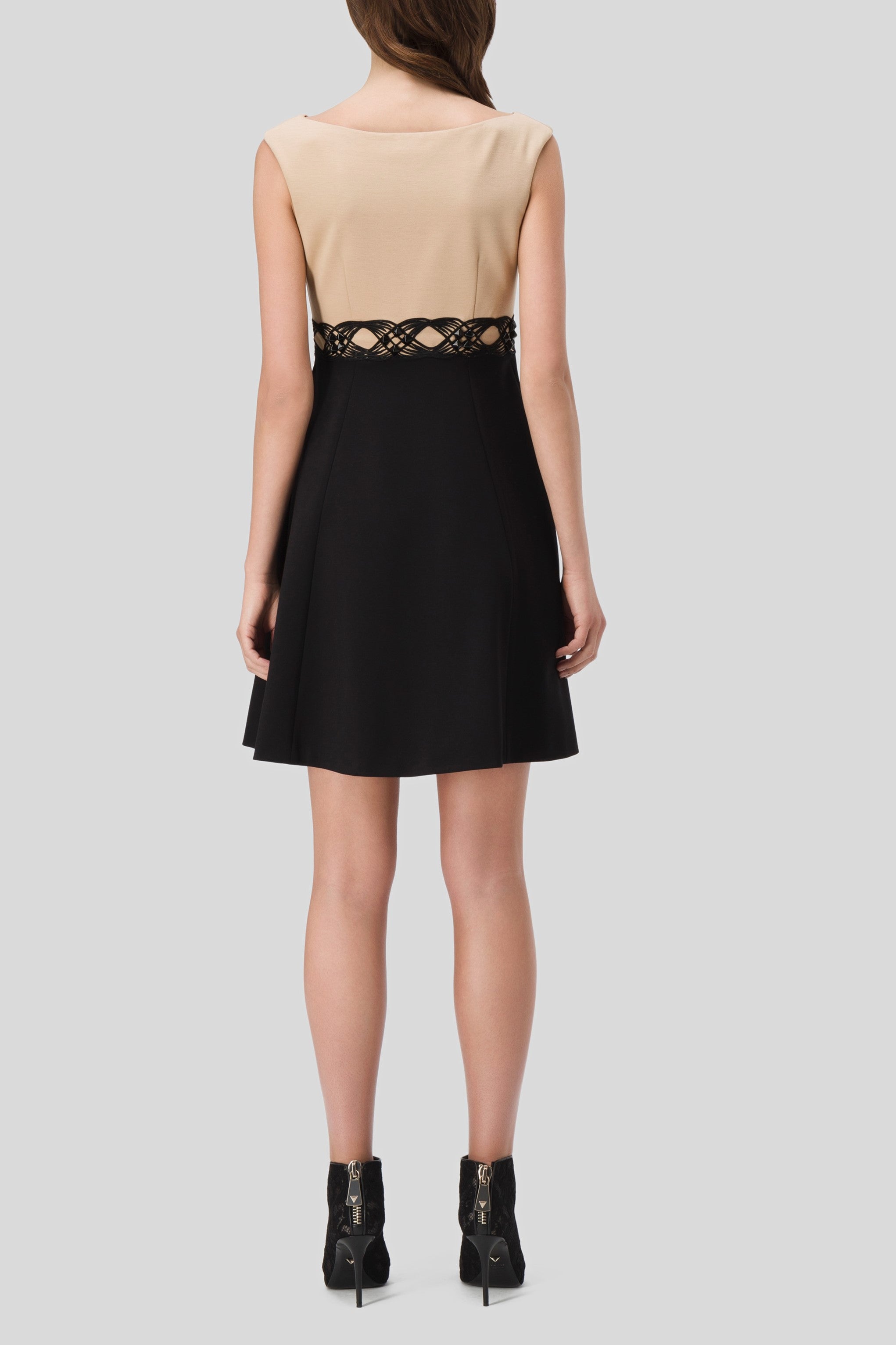 A woman wearing the Antonio Croce Annely Dress with Layered Design, featuring a beige top and black skirt, stands with her back to the camera. The A-line silhouette highlights a contrasting lace detail at the waist. She pairs the dress with black ankle boots.