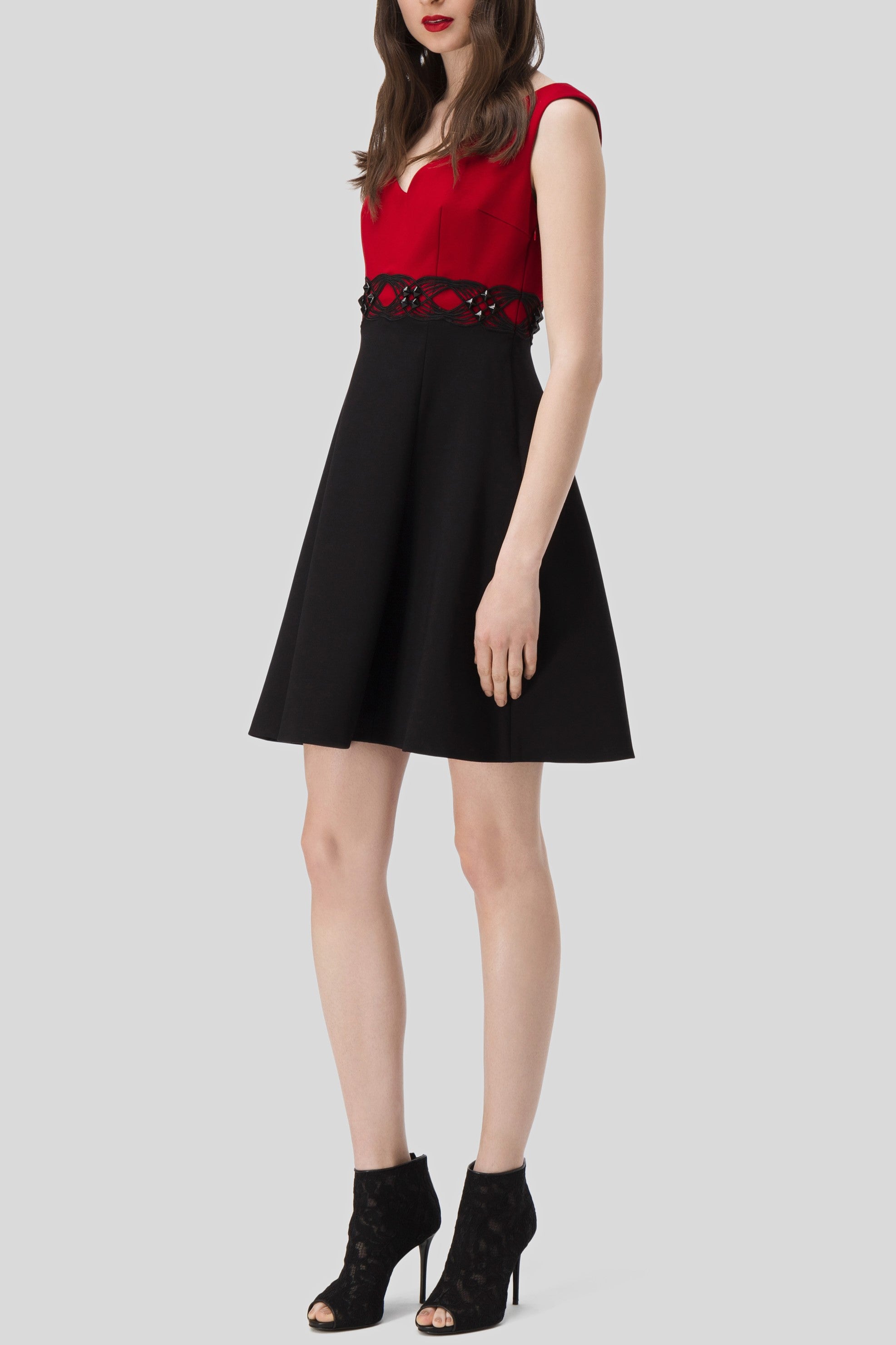 A woman stands facing forward, wearing the Antonio Croce Annely Dress with Layered Design, featuring a red and black honeycomb pattern at the waist. She pairs it with black open-toed heels. This knee-length, fitted dress flares out at the skirt, creating a timeless look against the plain gray background.