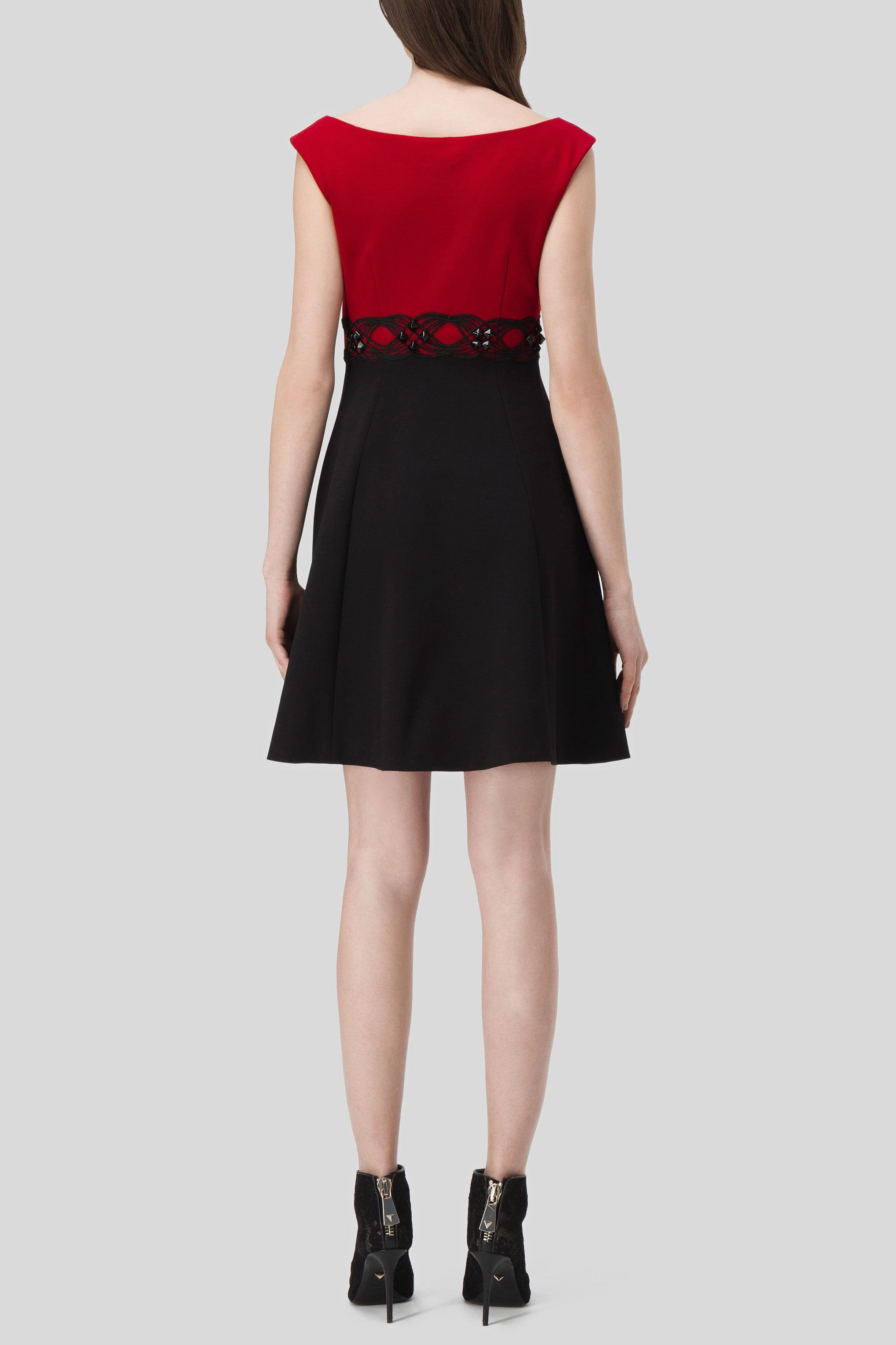 A woman facing away, wearing the Antonio Croce Annely Dress with Layered Design. The elegant sleeveless dress showcases a red top with a black A-line silhouette skirt, accented by a decorative lattice pattern at the waist. She completes her look with black ankle boots against a light gray background.