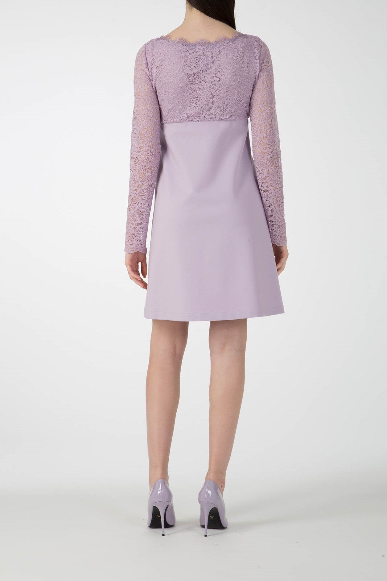 The woman is modeling the Antonio Croce Ardine Two-Tone Lace Dress, featuring vintage lace detailing on the sleeves in a romantic lavender hue. She stands against a plain white background, paired with matching lavender heels.