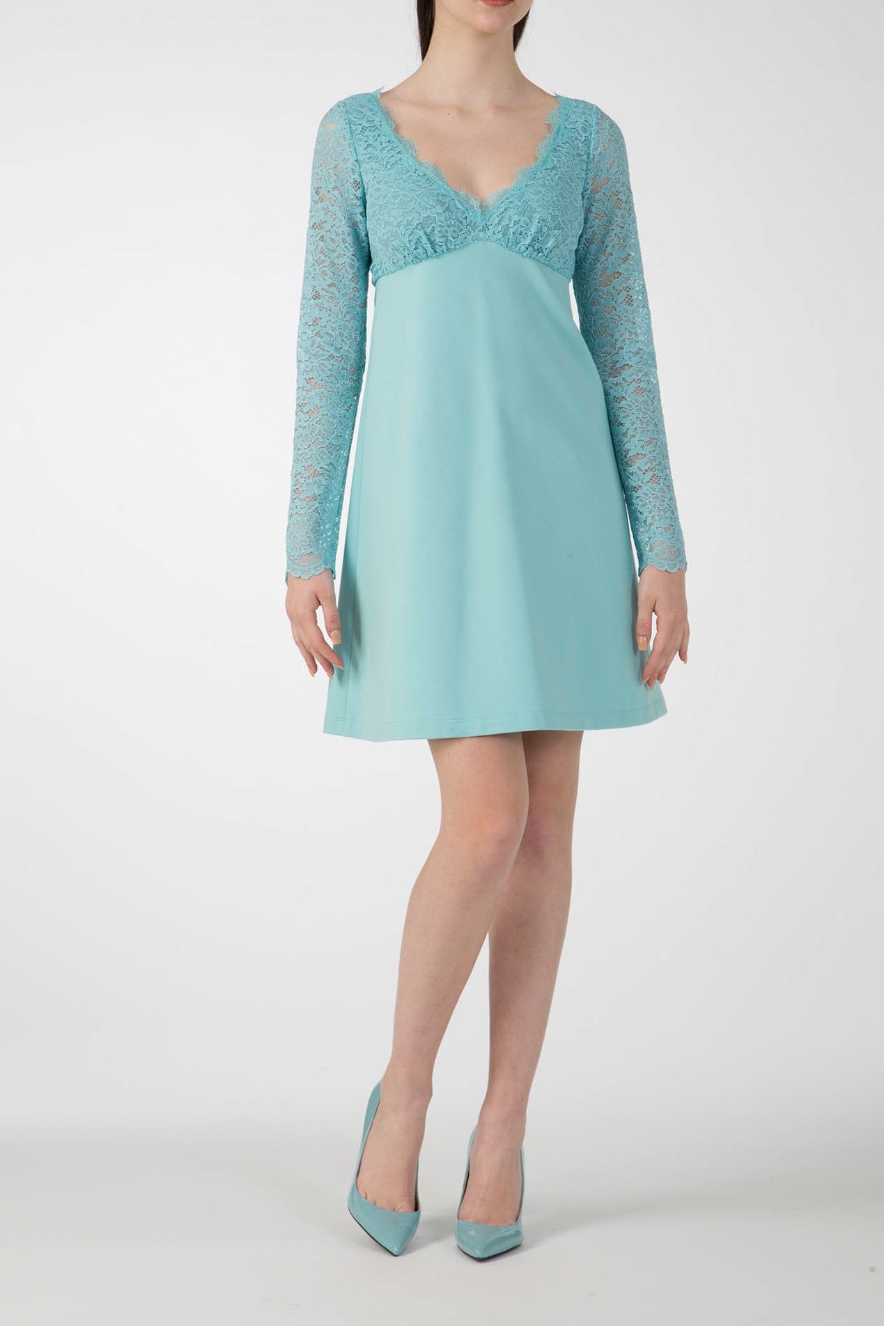 A woman stands against a plain backdrop, elegantly wearing the Antonio Croce Ardine Two-Tone Lace Dress in a romantic teal hue, with lace detailing on the sleeves. She complements it perfectly with matching teal high heels, while her face remains hidden from view.