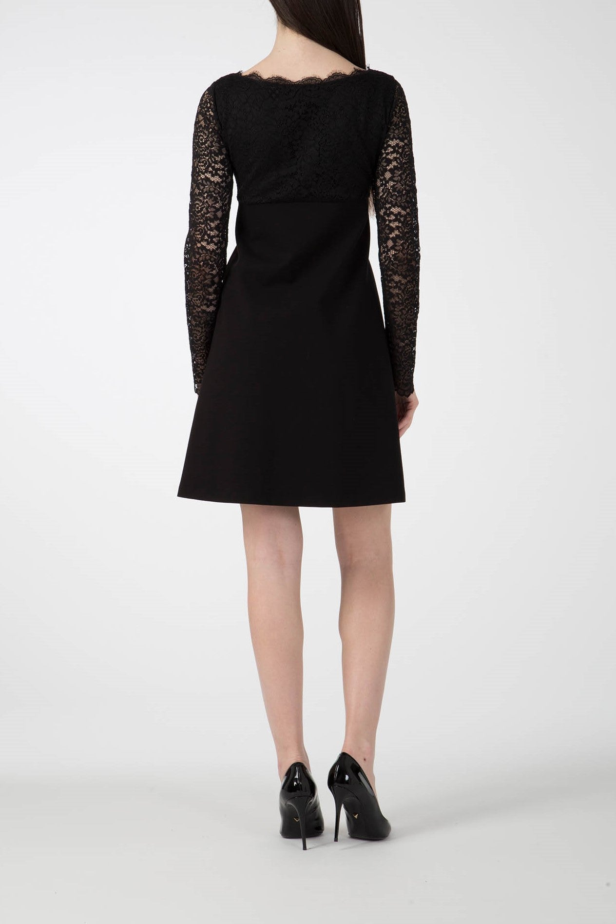 A person wearing the Antonio Croce Ardine Two-Tone Lace Dress, with its romantic lace detailing on the sleeves, and black high heels stands with their back to the camera against a neutral background.