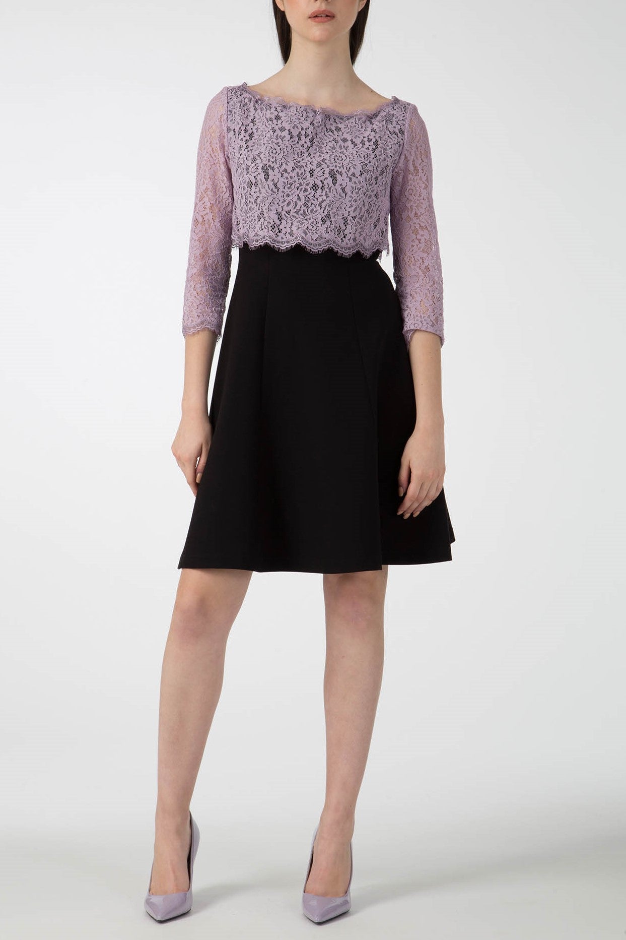 A woman in an Antonio Croce Artemide Two-Tone Dress, featuring a lilac lace bodice and black skirt, stands against a plain background. She completes her chic ensemble with matching lilac high heels.