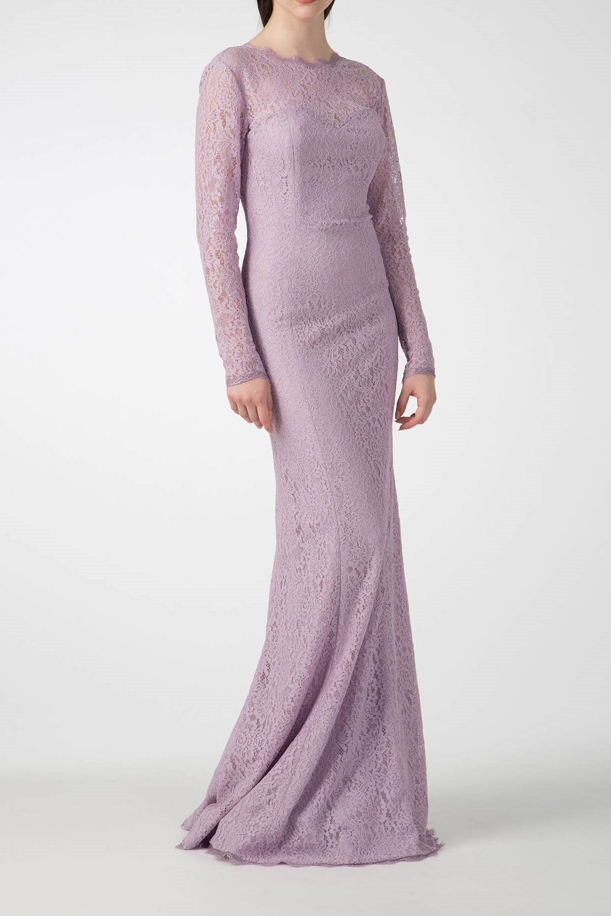 A person is elegantly adorned in a vintage piece—the Antonio Croce Australia Allover Lace Gown with a delicate light purple hue, featuring an illusion neckline, long sleeves, and a plunging back—poised gracefully against a plain background.