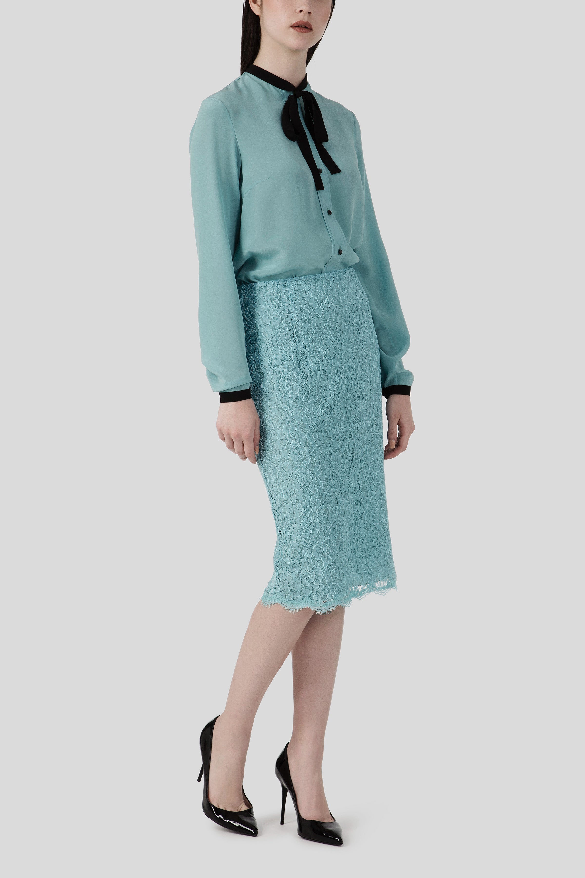 A woman stands wearing the Antonio Croce Barbie Elegant Blouse, a light blue creation crafted from sheer fabric and adorned with a black bow tie. She complements it with a matching lace pencil skirt and black high heels, exuding feminine style against the plain light gray background.