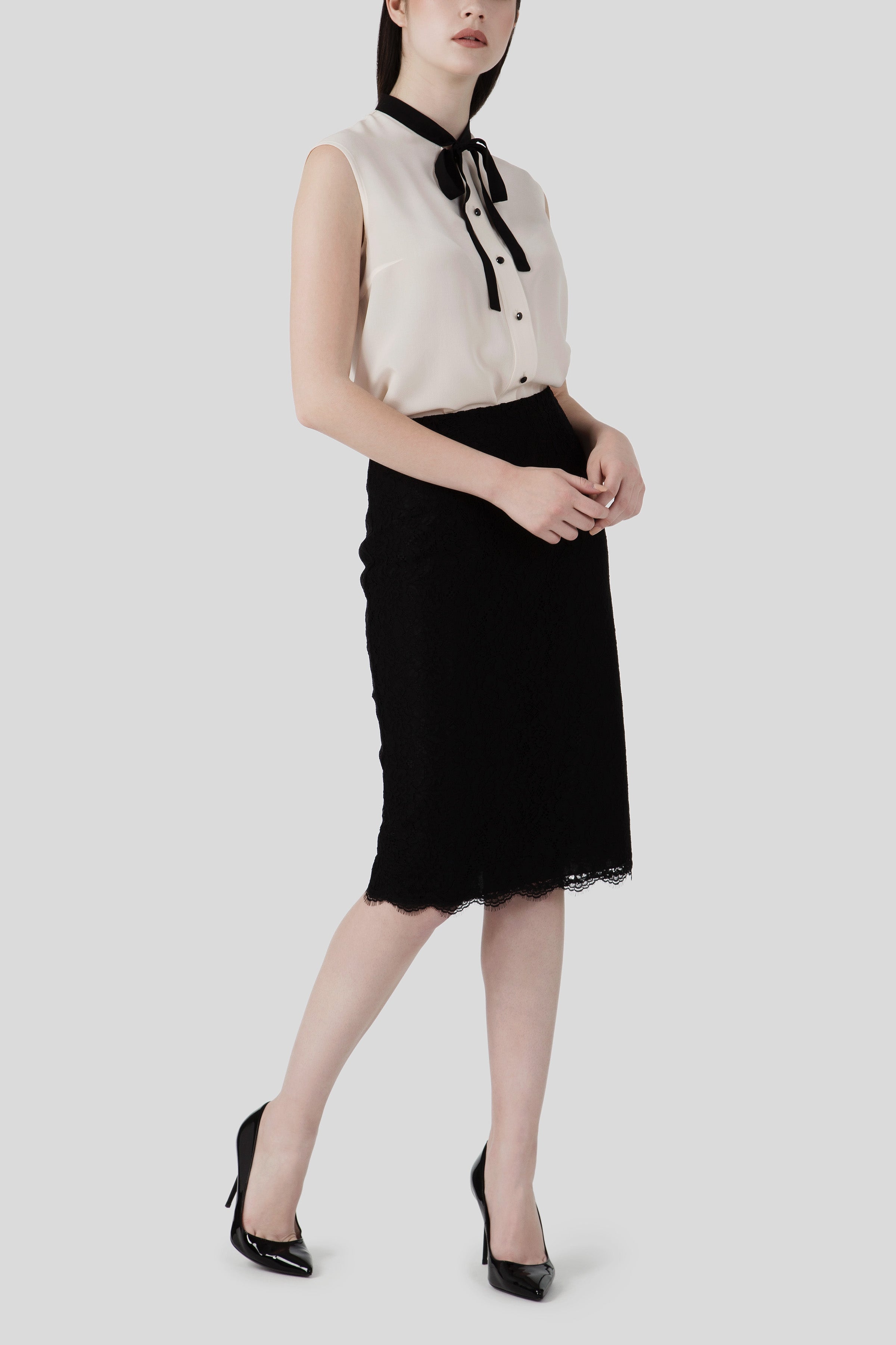 A woman radiates an effortlessly chic vibe in the Antonio Croce Bibi Flowy Blouse with Tie Neck, a sleeveless white top paired with a black lace pencil skirt. The blouse's sheer fabric adds an elegant touch to the ensemble. She completes her look with black heels, standing against a plain gray background.