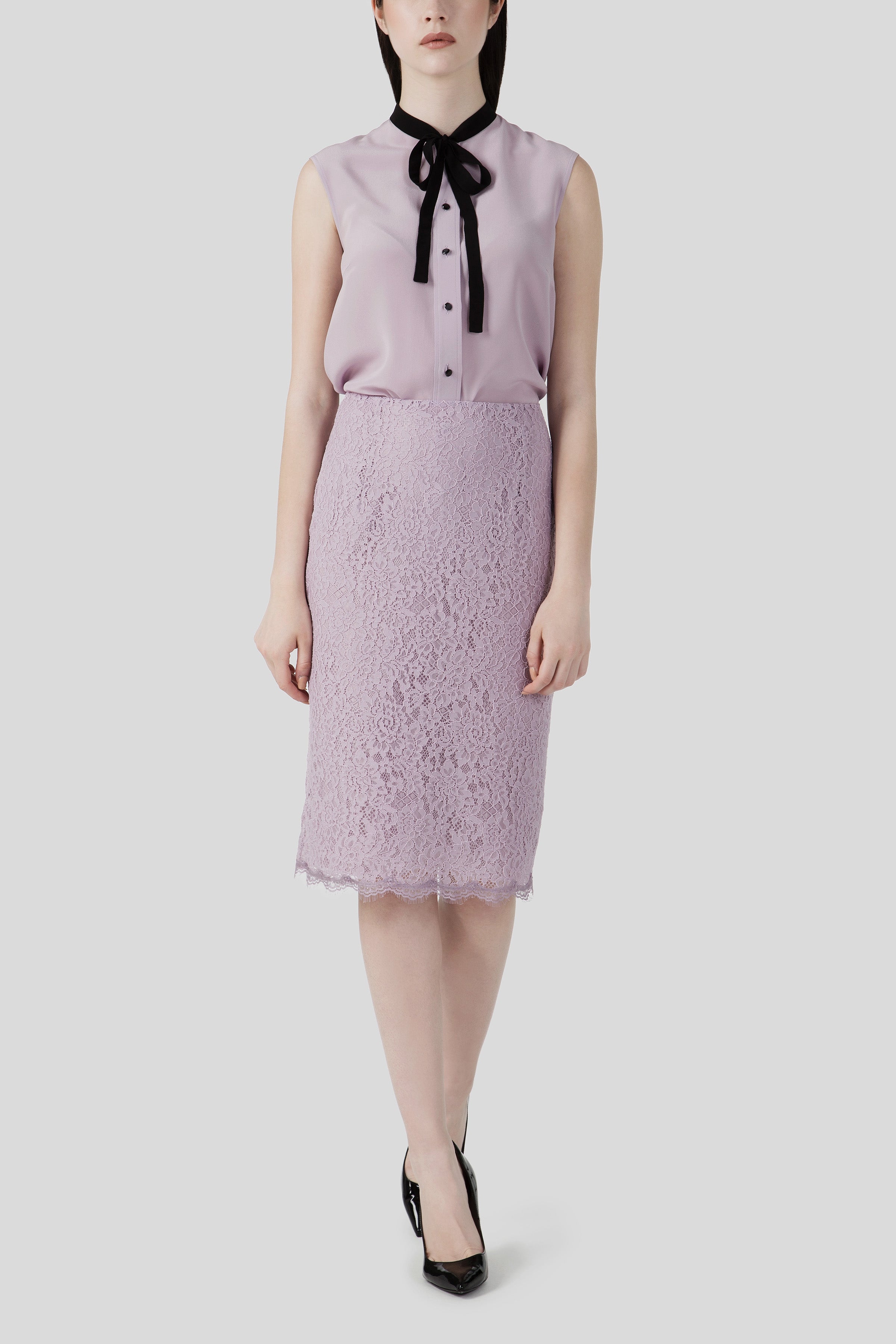 A person wearing the Antonio Croce Bibi Flowy Blouse with Tie Neck, styled sleeveless in a lavender hue and paired with a matching lace skirt, stands against a plain background. Their outfit, crafted from sheer fabric by Antonio Croce, is complemented by black heels.