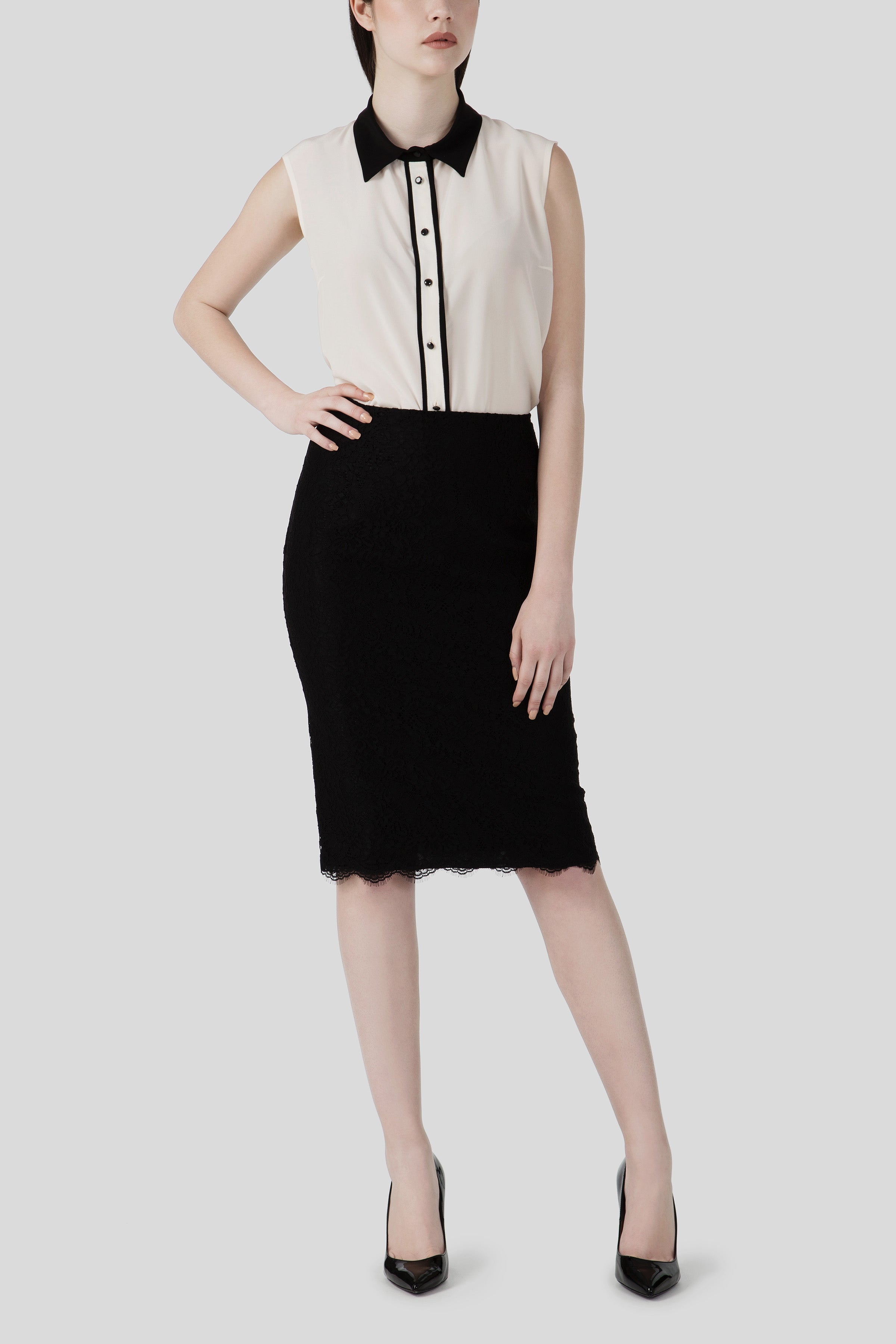 A person is wearing an Antonio Croce Brandy Sleeveless Shirt, featuring a contrasting black collar and buttons, paired with a black pencil skirt. They stand with one hand on their hip, adorned in vintage-inspired black high-heeled shoes against a light gray background.