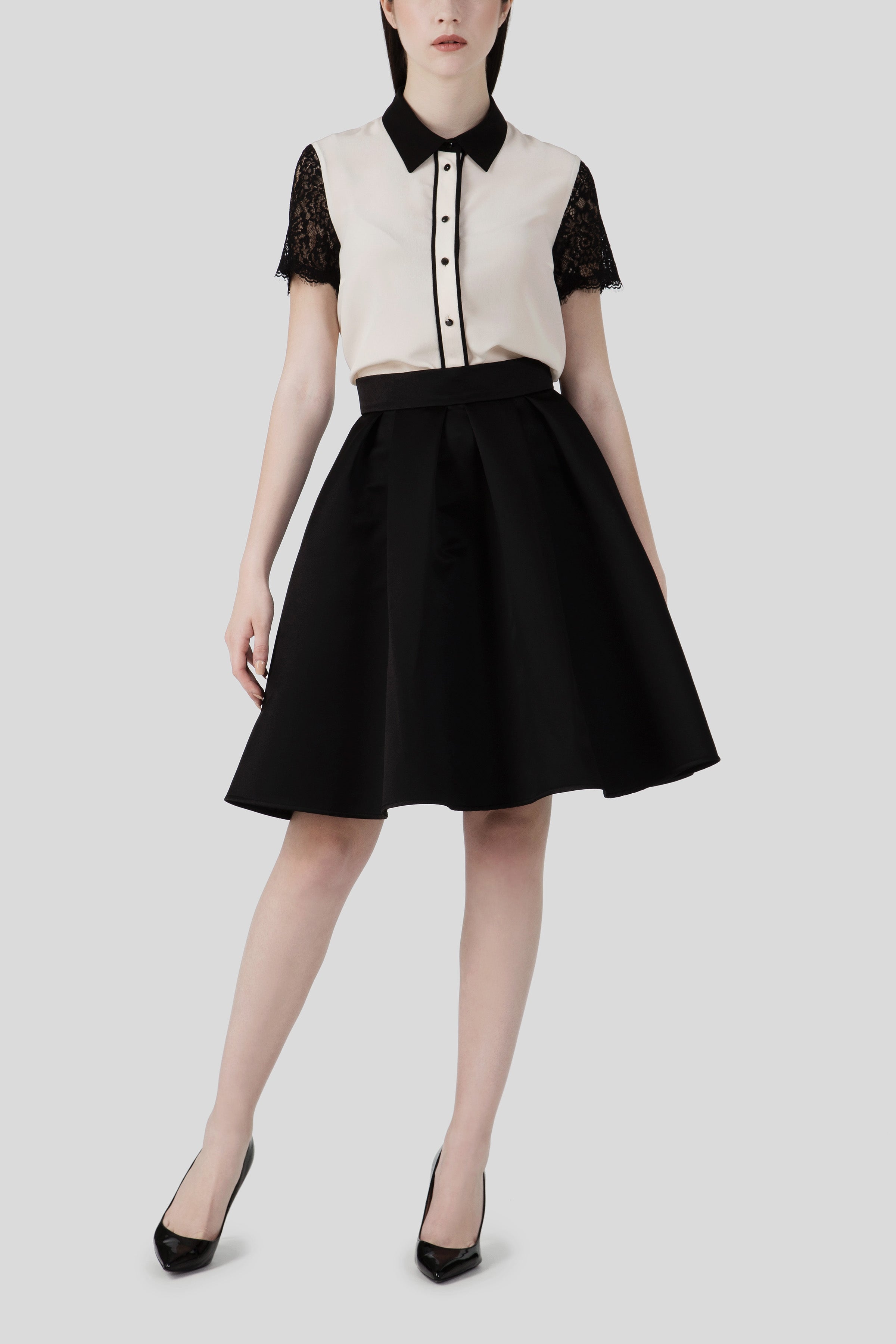 Someone dressed in a black and white dress featuring lace sleeves and a flattering silhouette, complemented by the Antonio Croce Shelby A-Line Skirt. The ensemble is completed with black heels and set against a plain gray background.