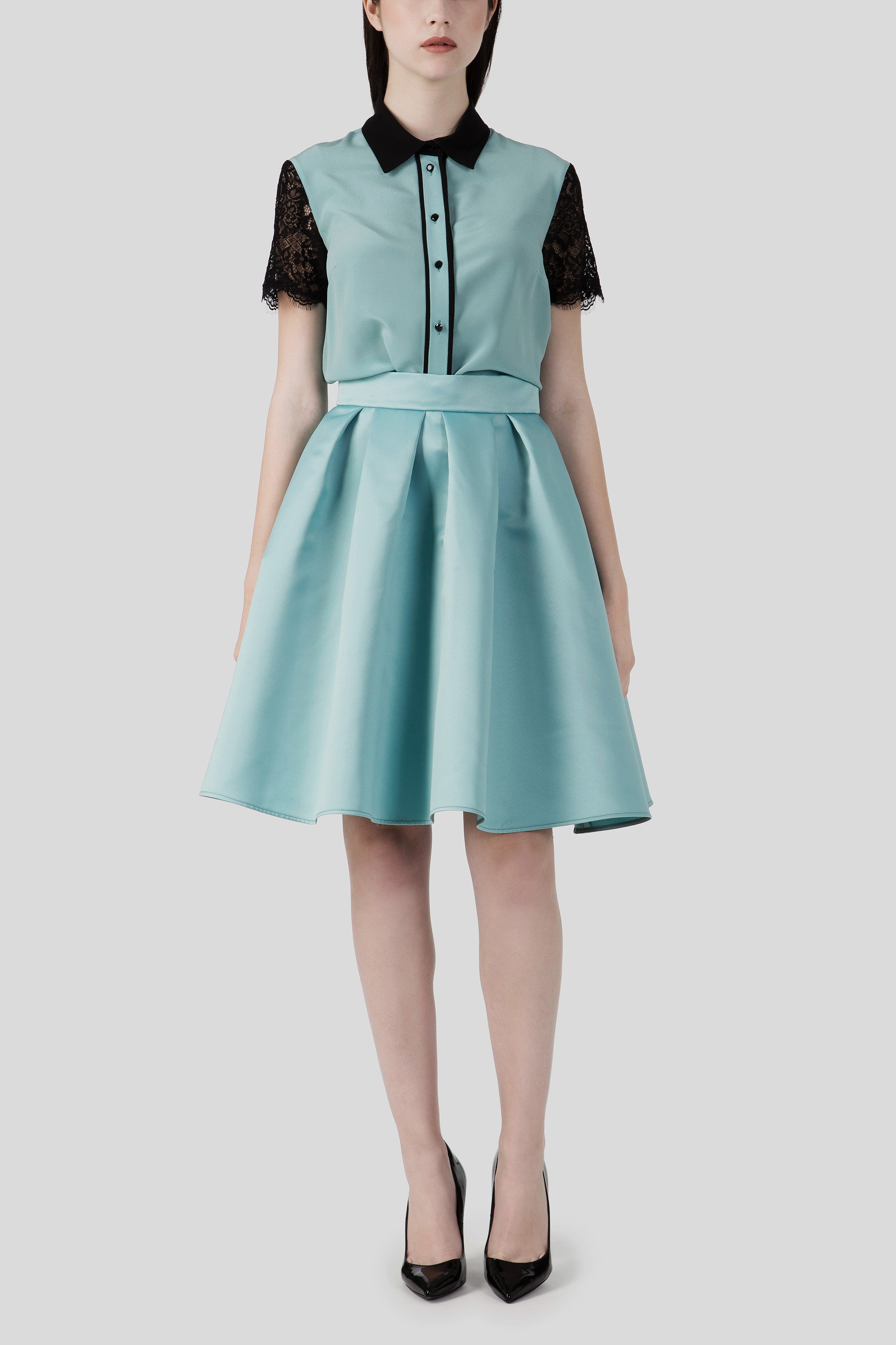 A woman in an Antonio Croce Shelby A-Line Skirt, featuring a teal color with black lace sleeves and collar for a flattering silhouette, stands against a plain background. This vintage piece is elegantly completed with black high heels from the brand Antonio Croce.