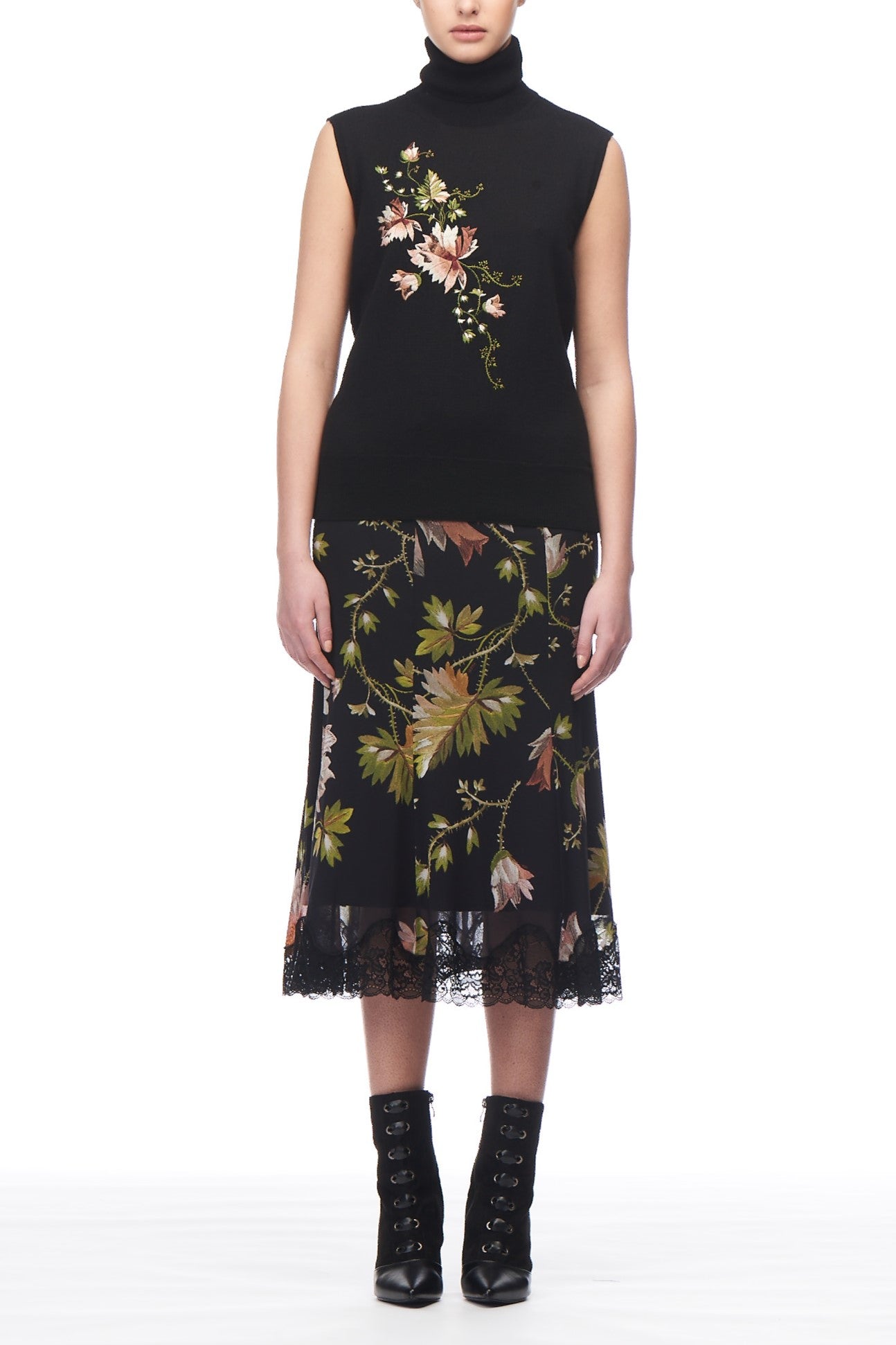 A person wearing a Fuzzi Woman Embroidered Top in black, adorned with floral designs, complemented by a vibrant pink floral skirt featuring lace trim. The ensemble is completed with black lace-up ankle boots and set against a plain white background.