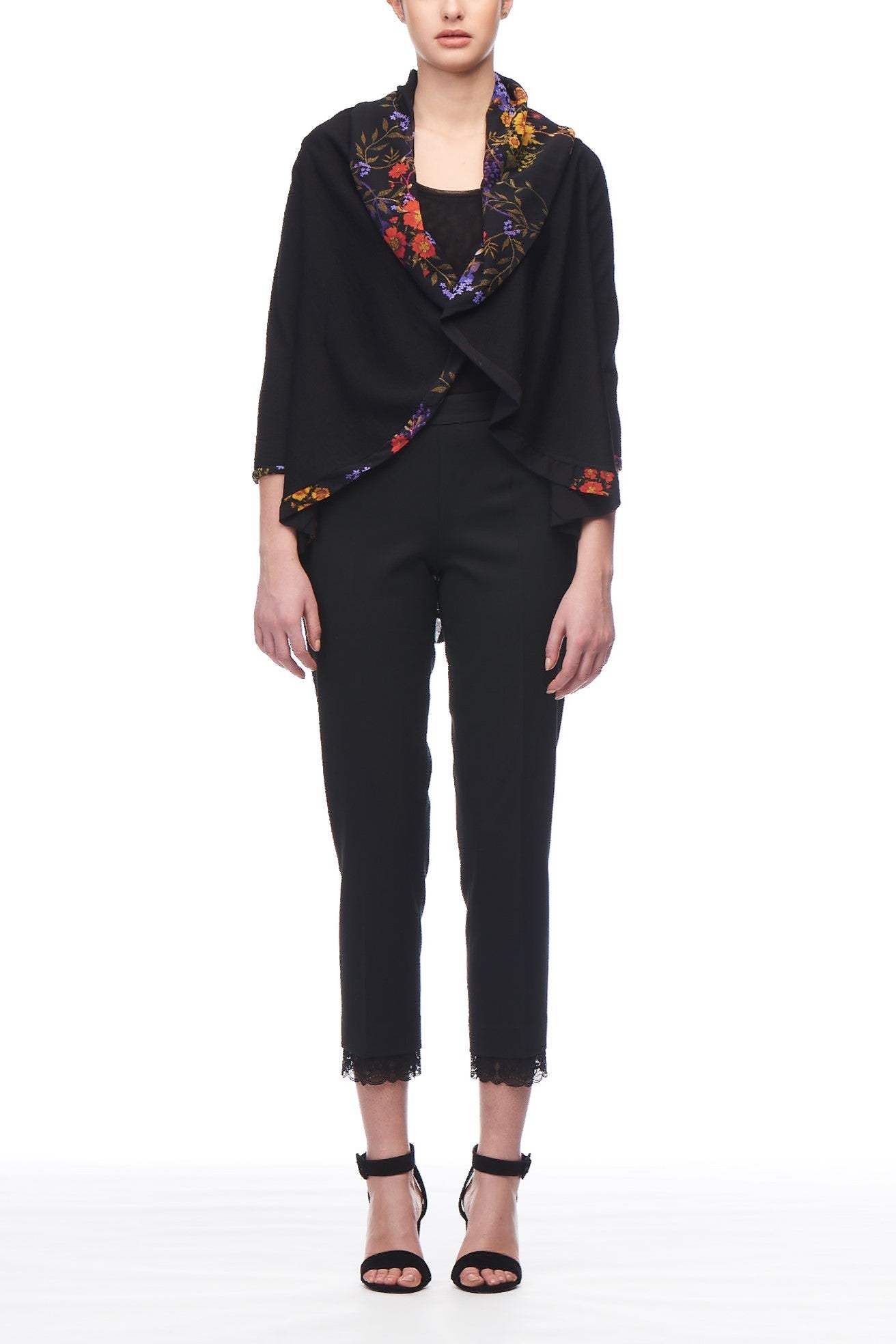 A person wearing a Fuzzi Woman Black Cardigan featuring vibrant floral accents over a black top, paired with black pants and high-heeled sandals. The colorful embroidery stands out against the plain white background, adding character to the ensemble.