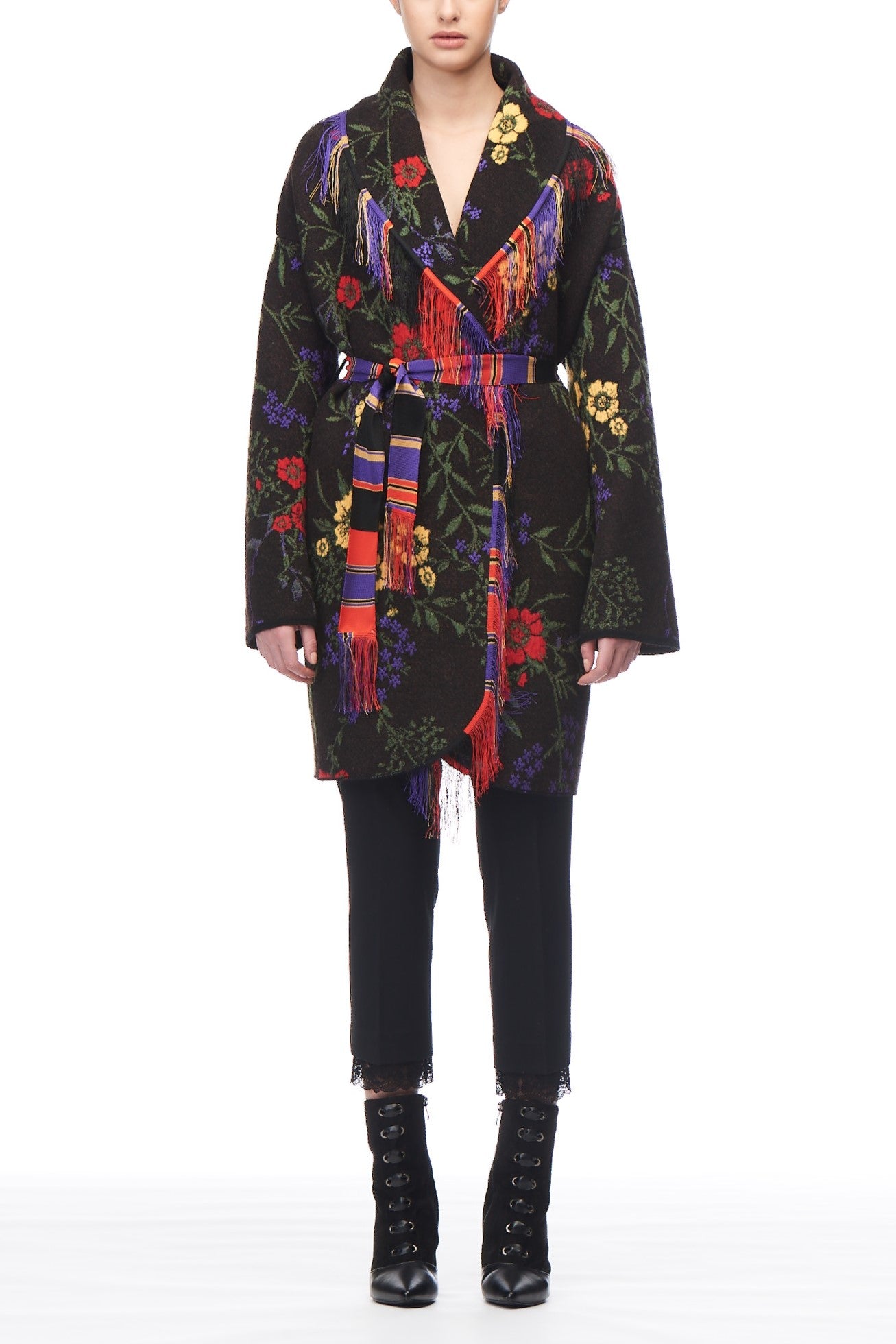 A person wearing the chic Fuzzi Woman Floral Wrap Coat with colorful floral patterns and a vibrant multicolored striped belt. They complete the look with black pants and lace-up boots against a pristine white background.