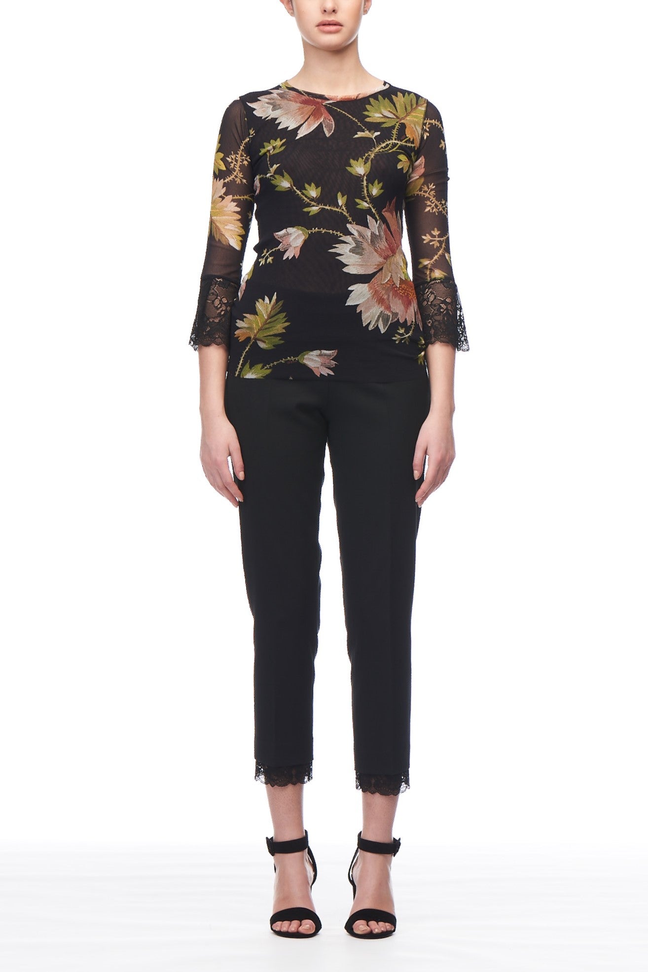 A person wearing the Fuzzi Woman Floral T-Shirt, featuring vintage sheer black fabric with floral designs and lace sleeves, paired with black pants and ankle-strap heels. The outfit showcases large floral patterns in earthy tones, highlighted against a white background.