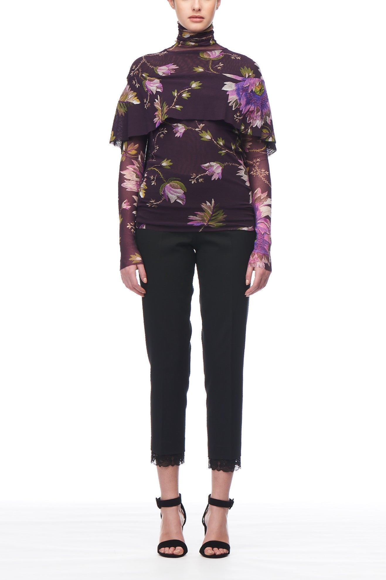 A person stands against a white background wearing the Fuzzi Woman Ruffled Floral Mesh Top, featuring a dark floral print and layered short sleeves, paired with black cropped pants and black high-heeled sandals. The outfit displays purple and green flowers while their hands rest at their sides.