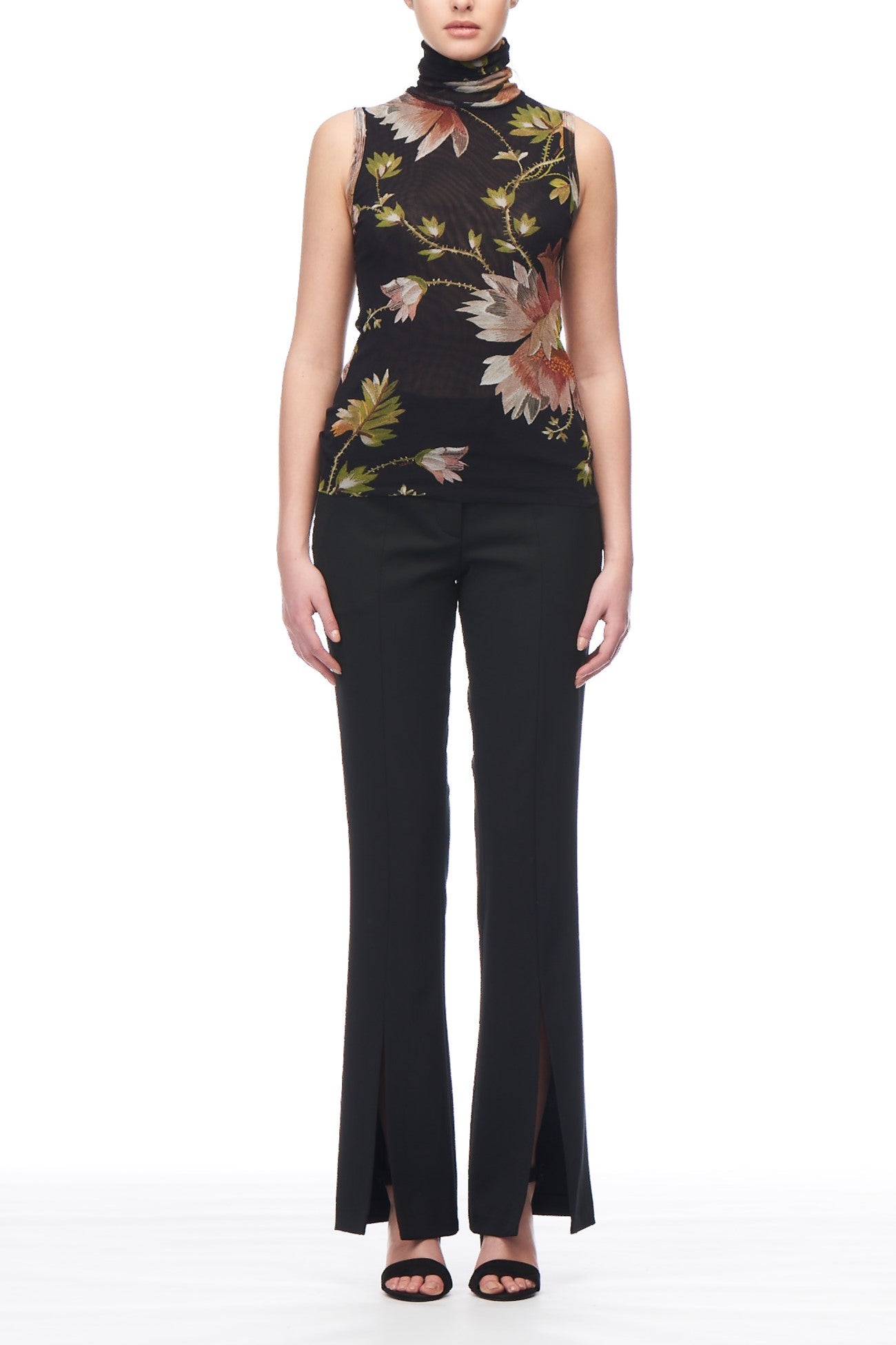 A person wearing the Fuzzi Woman Floral Mock Neck Top, featuring a sleeveless design and floral patterns on a black background, paired with black pants with ankle slits, is standing against a white backdrop. Their look is completed with black open-toe shoes.