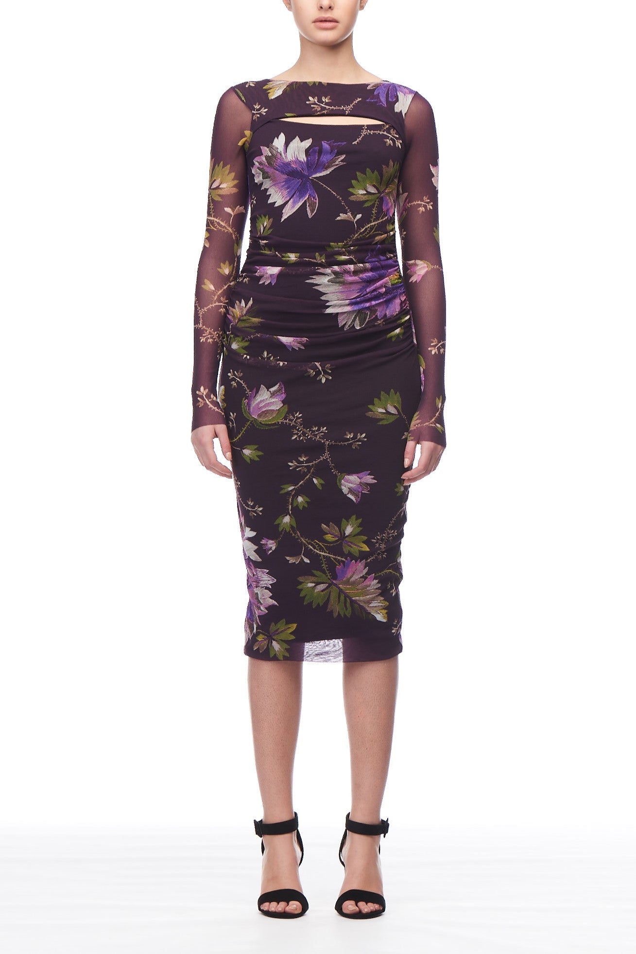 A person is dressed in a Fuzzi Woman Floral Mesh Midi Dress, boasting a dark floral print with purple and green blooms. The ruched design adds elegance, complemented by black strappy high-heeled sandals.