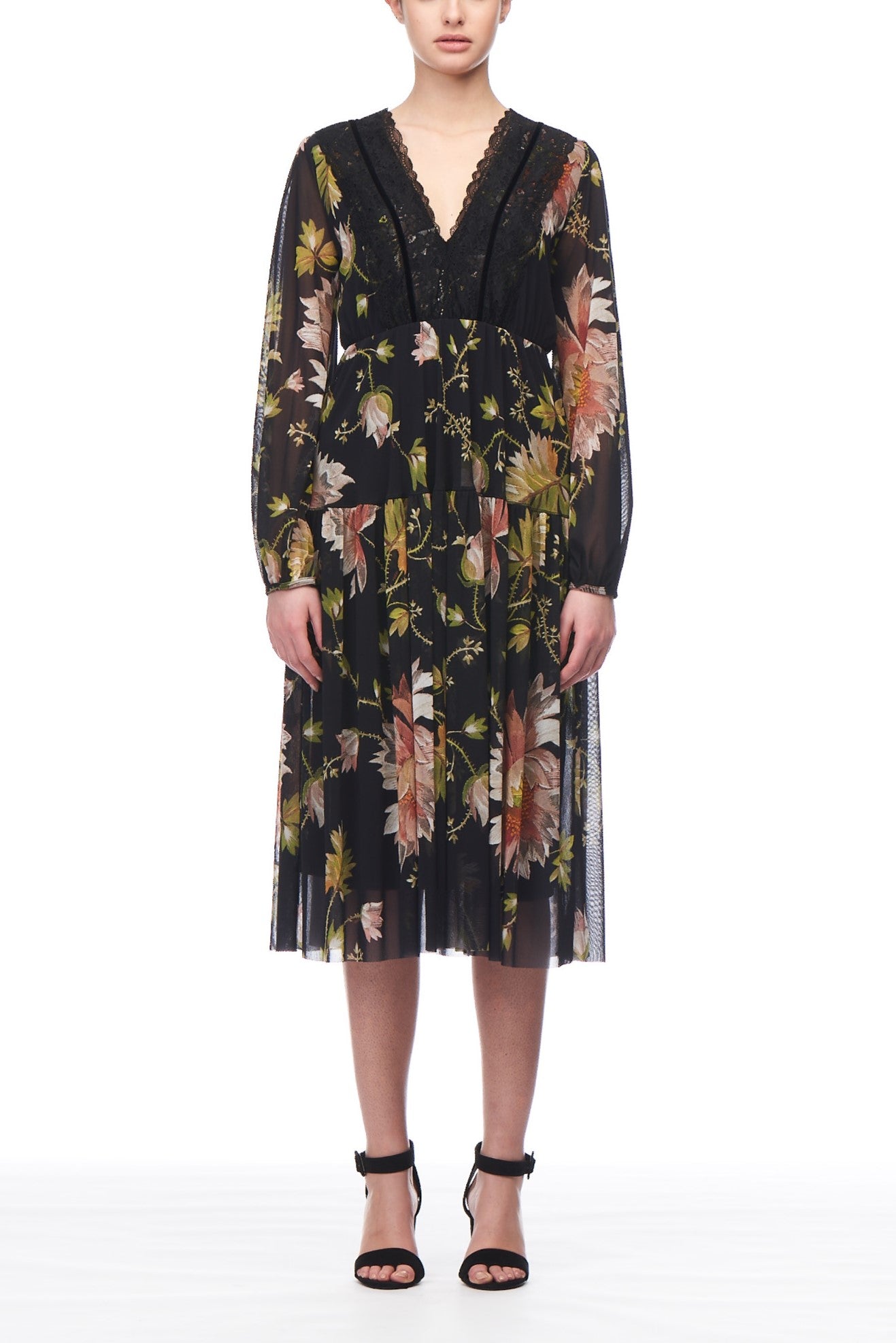 A person is wearing the Fuzzi Woman Short Mesh Dress by Fuzzi, a long-sleeved dress with a v-neckline and pleated skirt. The dress showcases sheer mesh fabric adorned with colorful floral patterns. They are also wearing black high-heeled sandals while standing against a plain white background.