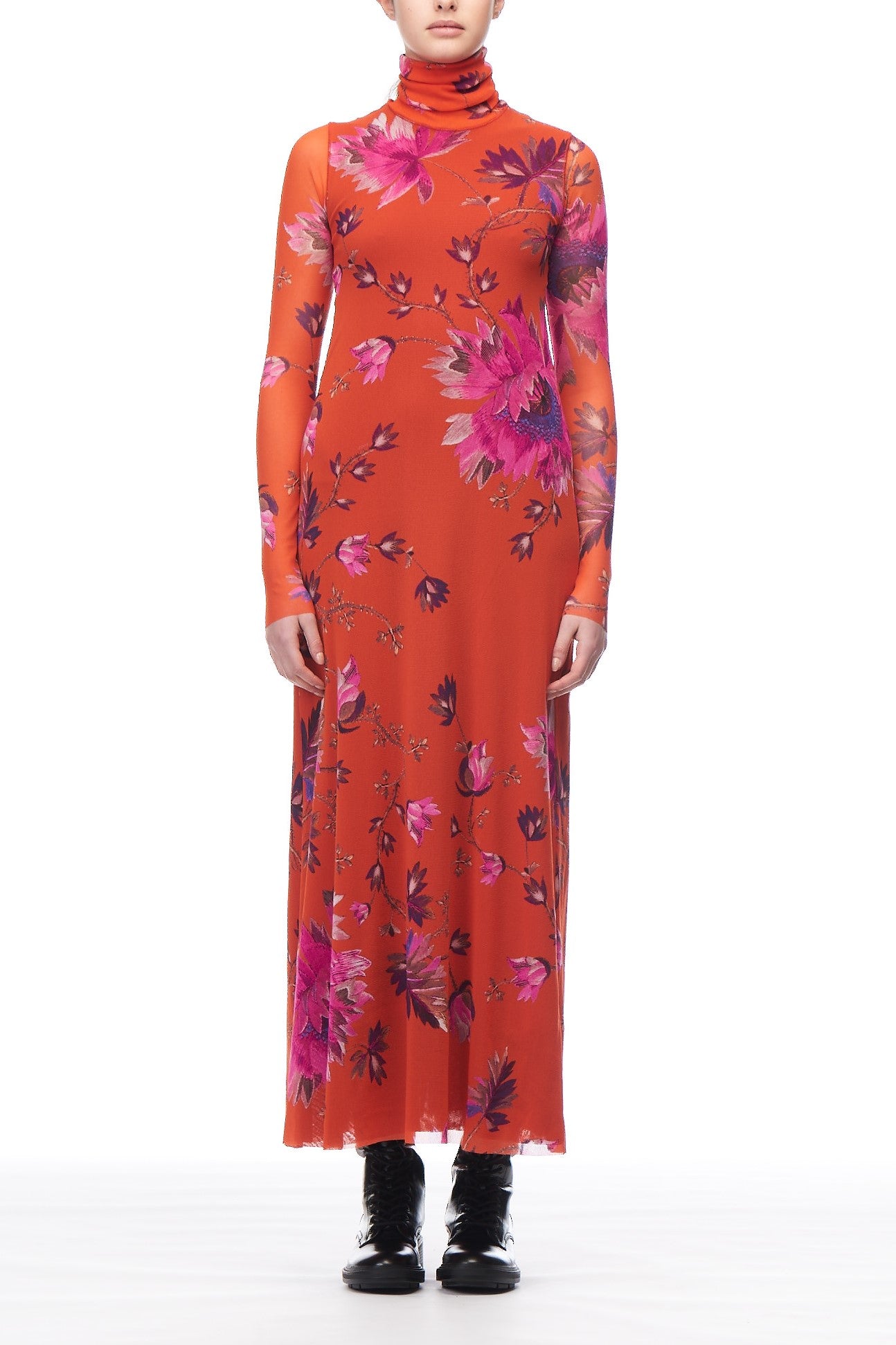 A person wearing the Fuzzi Woman Floral Mesh Maxi Dress, featuring a vibrant pink floral print, stands against a plain white background. The floor-length orange outfit is perfectly complemented by black ankle boots.