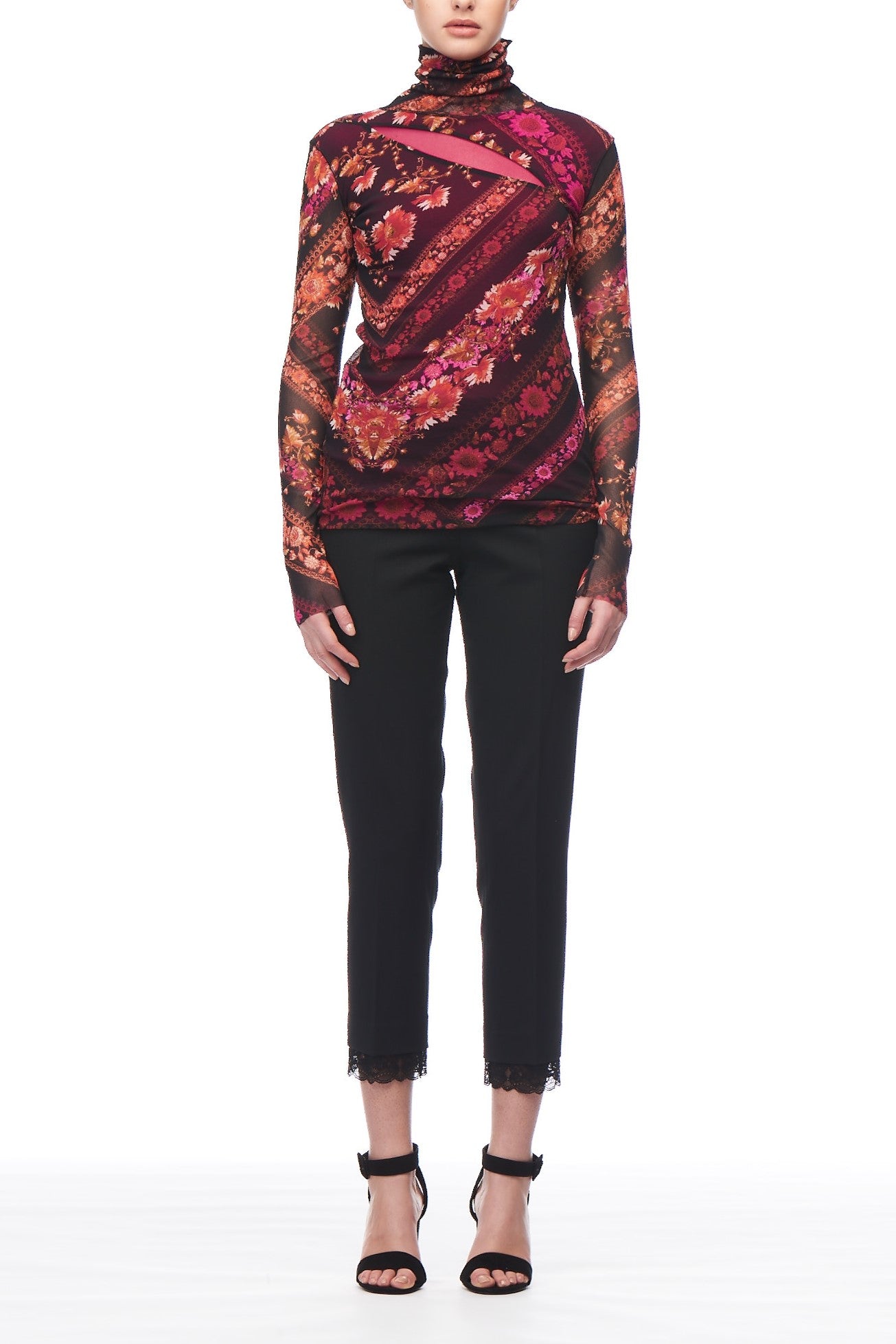 The individual is dressed in a Fuzzi Woman Floral Mesh Turtleneck from the brand Fuzzi, adorned with vibrant geometric patterns in shades of red, purple, and orange. Elegantly complemented by black pants featuring lace hems and black open-toed heels.